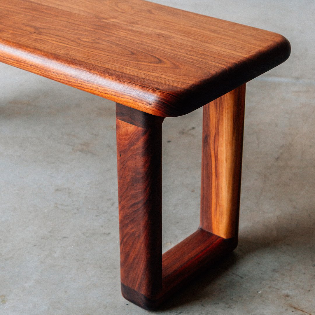 Close-up of a wooden table with a smooth, polished surface and rounded edges, showing the corner supported by a curved wooden leg.