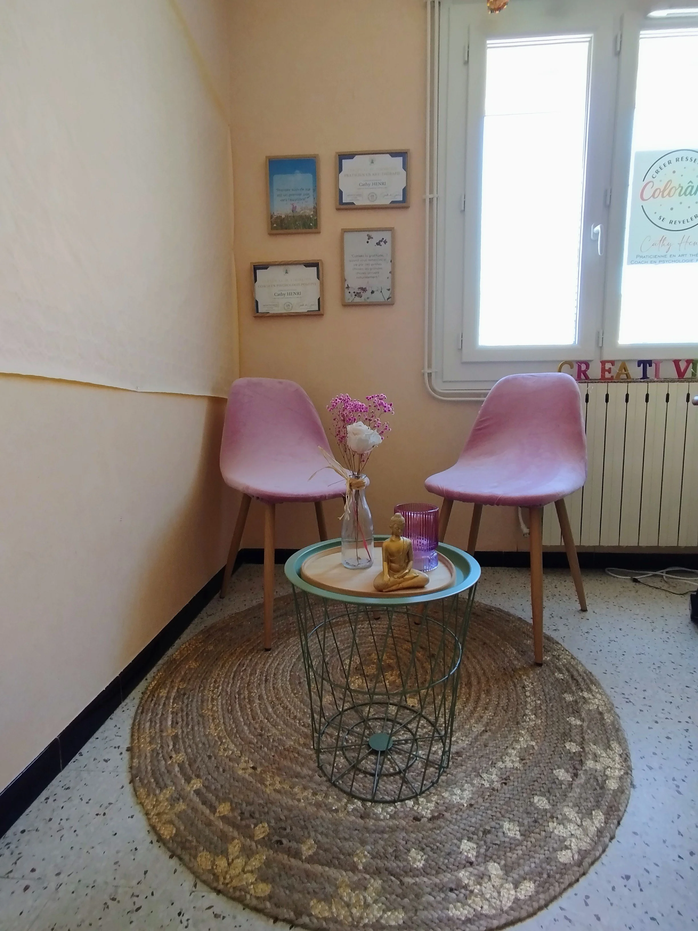 Un coin d'attente avec deux fauteuils roses, une petite table ronde en métal avec une bouteille contenant des fleurs, une statue de bouddha dorée et un vase violet. Un tapis rond en fibres naturelles est au sol, avec un mur décoré de diplômes et affiches, et une fenêtre laisse entrer de la lumière.