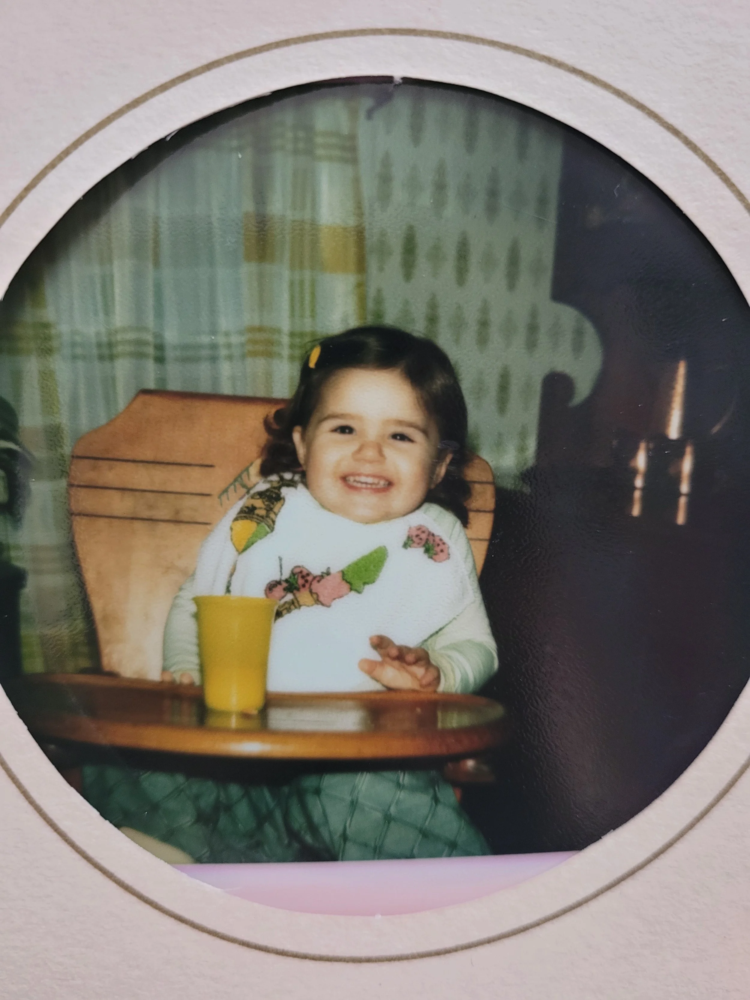 A young girl sitting at a wooden table with a yellow cup in front of her, smiling at the camera. She has curly dark hair with a yellow hair clip, and is wearing a colorful sweater with a collar. The background shows a curtain and a dark-colored object, possibly a piece of furniture or a fireplace.