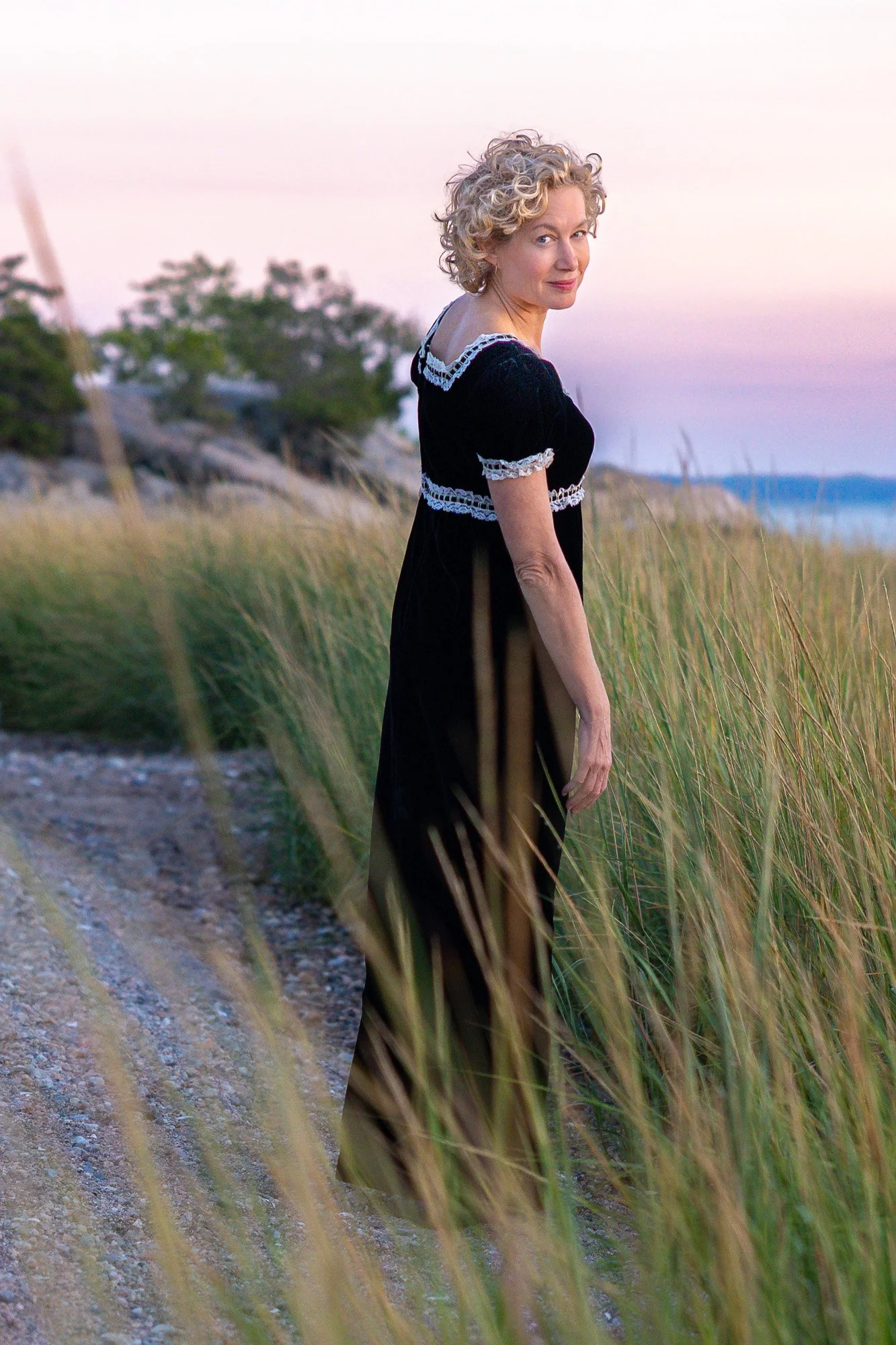 A woman in a black dress standing on a sandy path next to tall grass during sunset at the beach.
