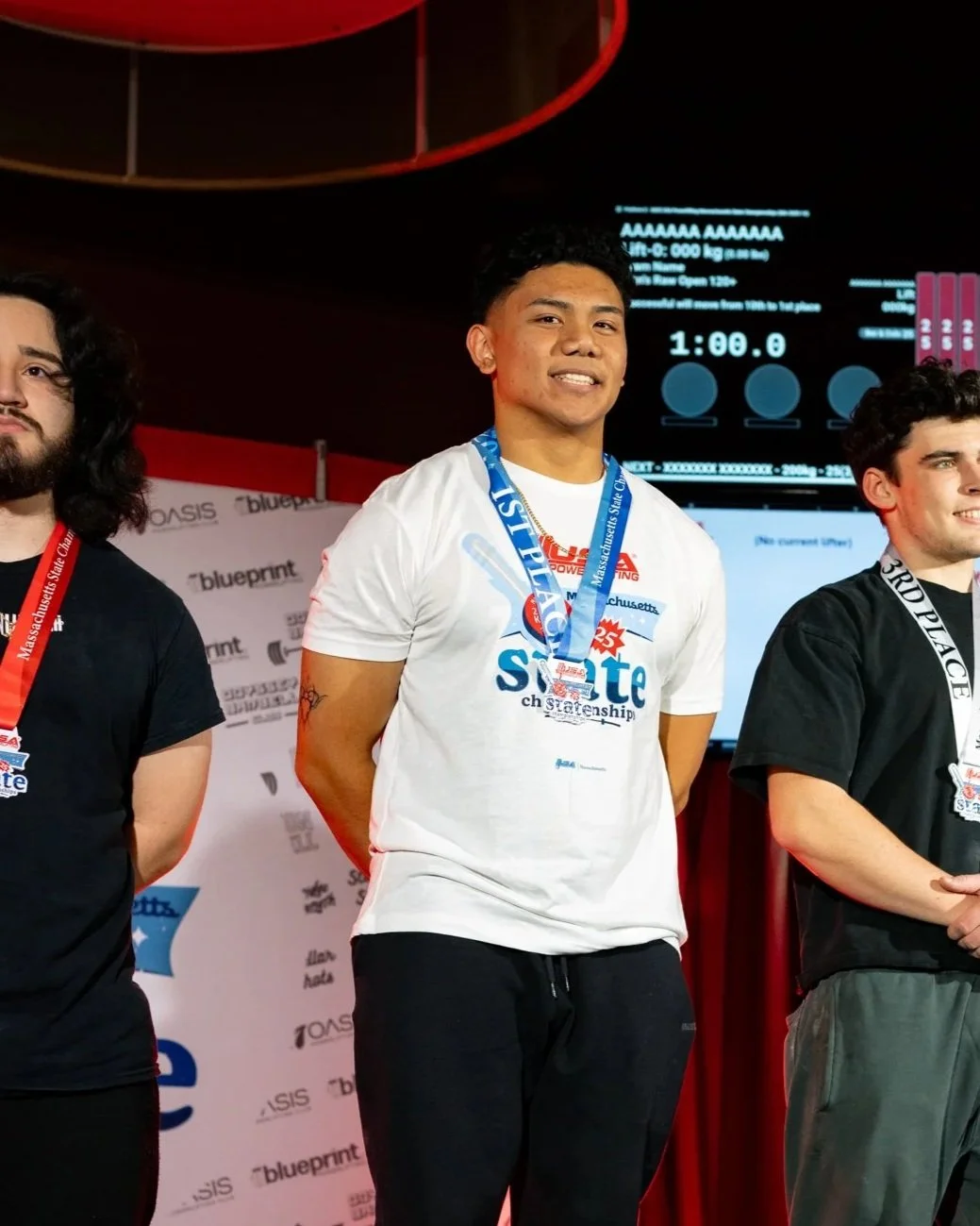 Three young men standing on a podium at a competition, wearing medals around their necks, with a backdrop displaying event details and a digital timer.