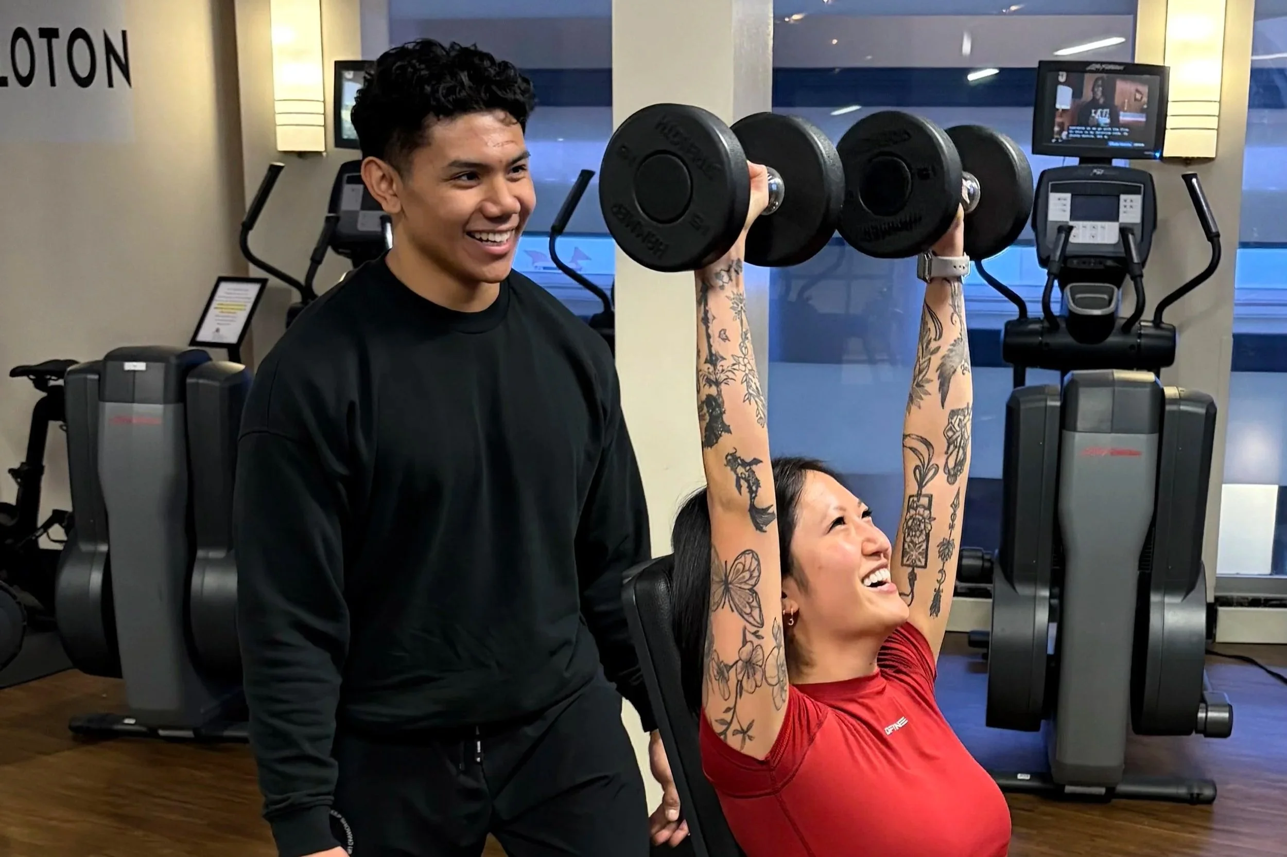 A woman with tattoos in a red shirt is lifting dumbbells while seated in a gym, smiling, with a trainer watching and smiling nearby.