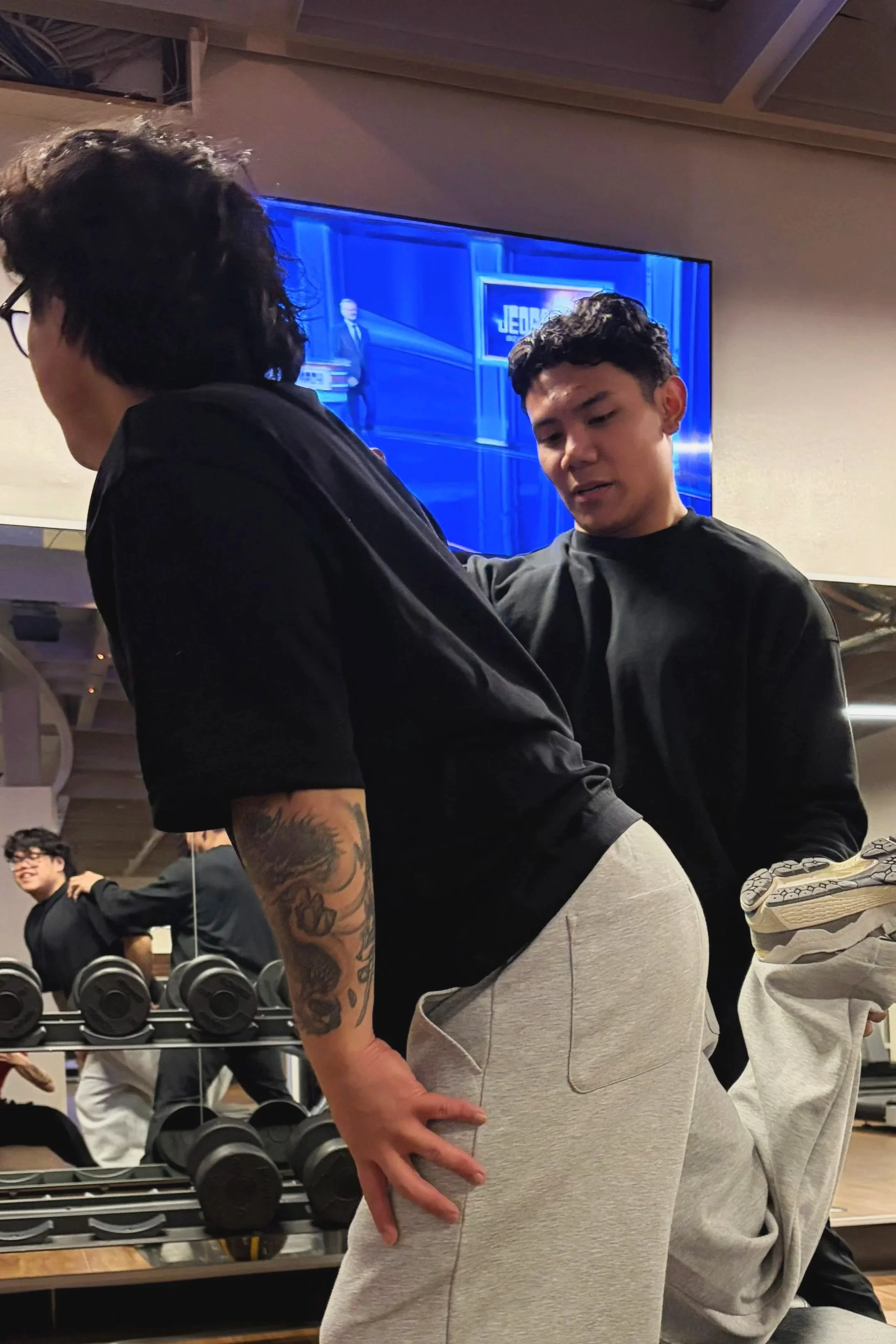 Two young men at a gym with a TV monitor in the background, one of them bending forward with his hand on his hip and the other assisting him, while another person is exercising in the background.