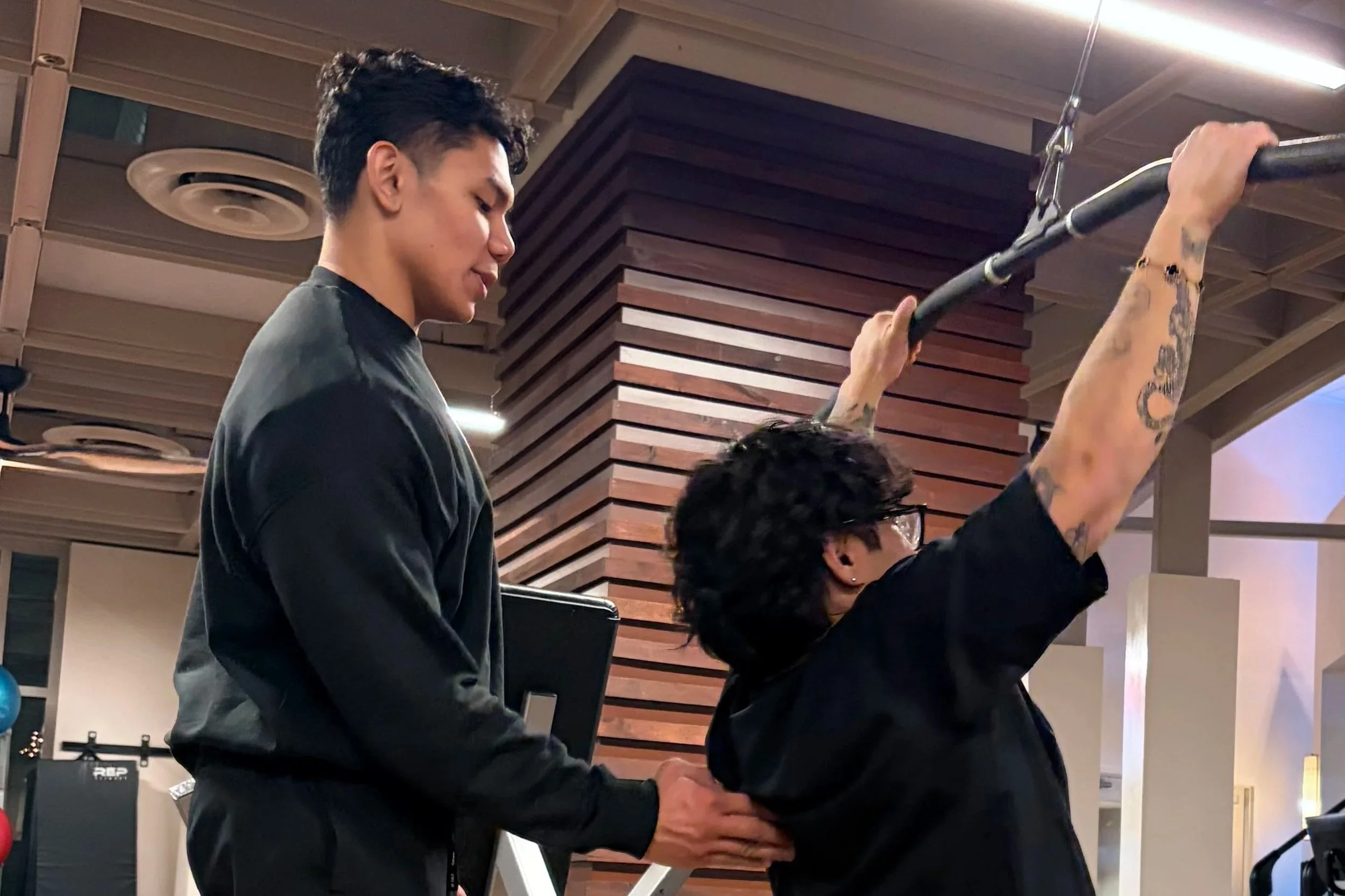 A personal trainer assisting a young man with a pull-up exercise in a gym.