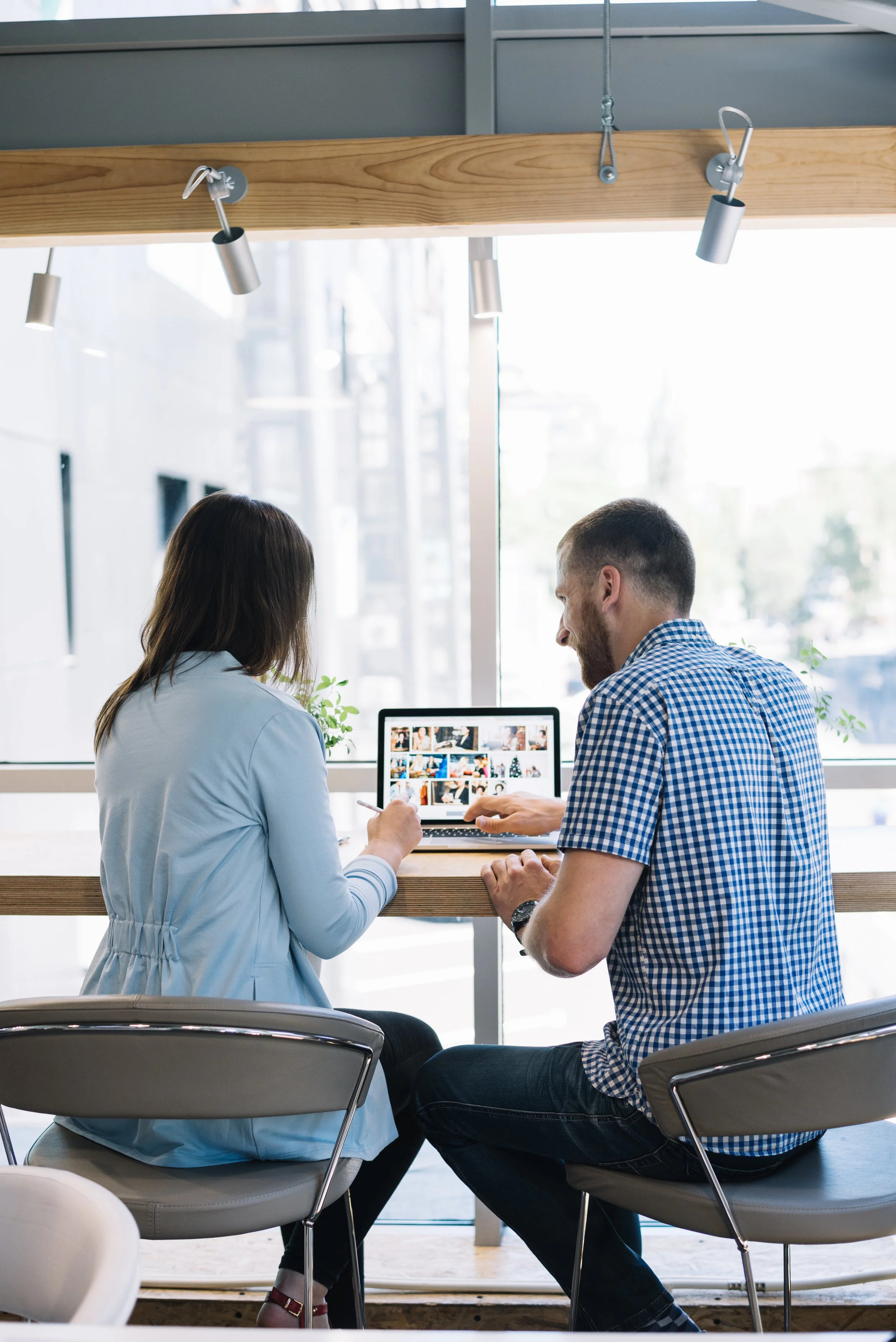 Dos personas sentadas en una mesa frente a una ventana, trabajando en una computadora portátil con una galería de fotos.