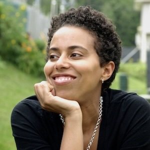 Close-up of Asha Noel-Hart with short curly hair, smiling, wearing a black top and silver necklace, outdoors in a park.