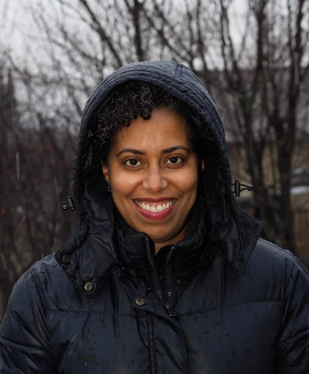 Asha Noel-Hart is smiling, wearing a black hooded jacket outdoors in rainy weather, with blurred trees in the background.