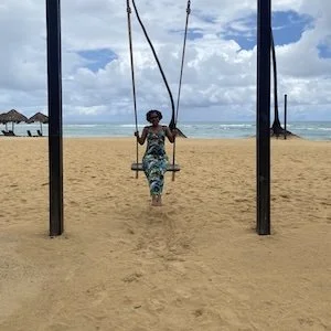 Person sitting on a swing set at the beach with ocean and cloudy sky in the background.