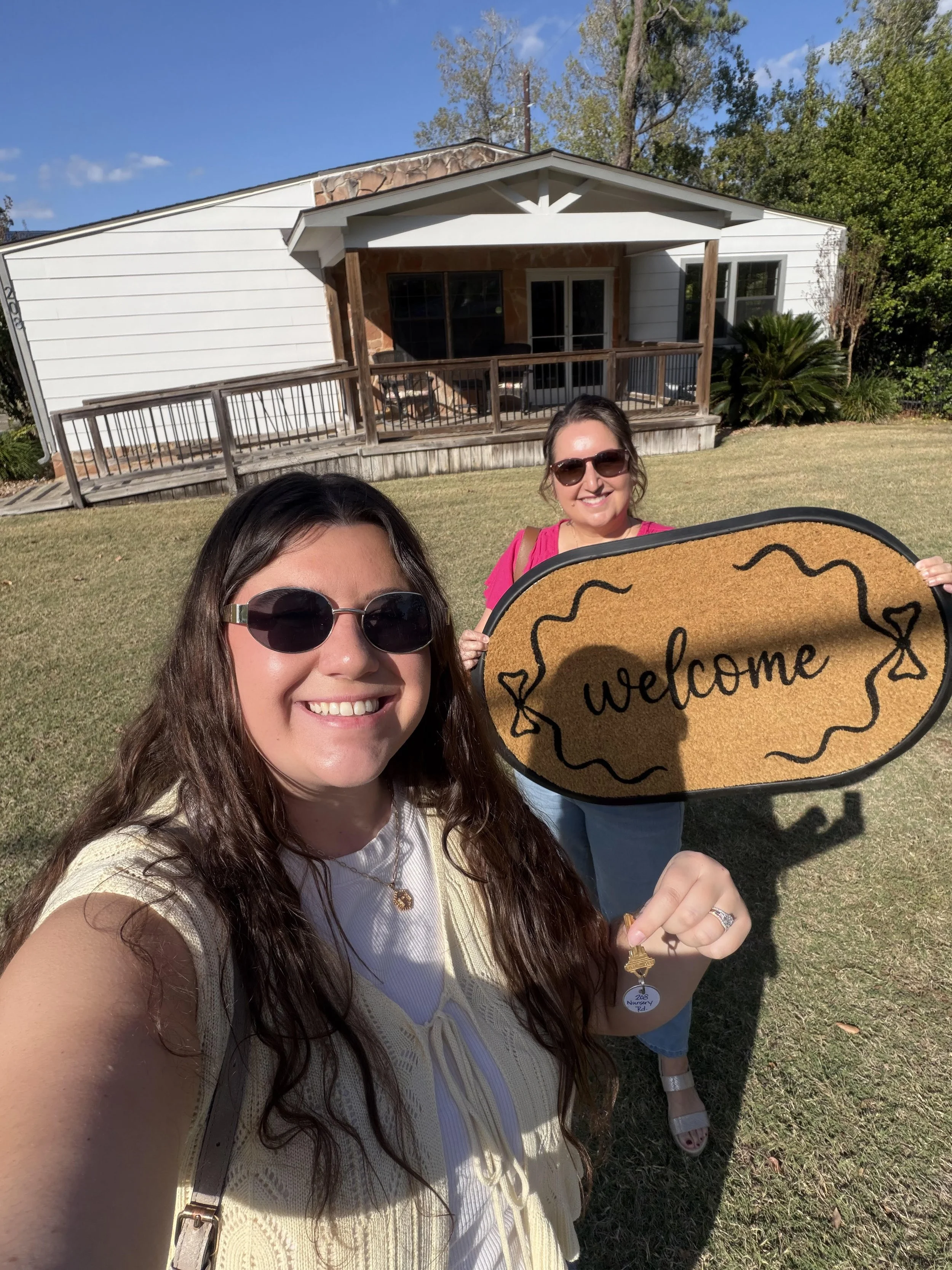 Two women smiling outside a house on a sunny day, one holding a 'welcome' sign, with a porch and trees in the background.
