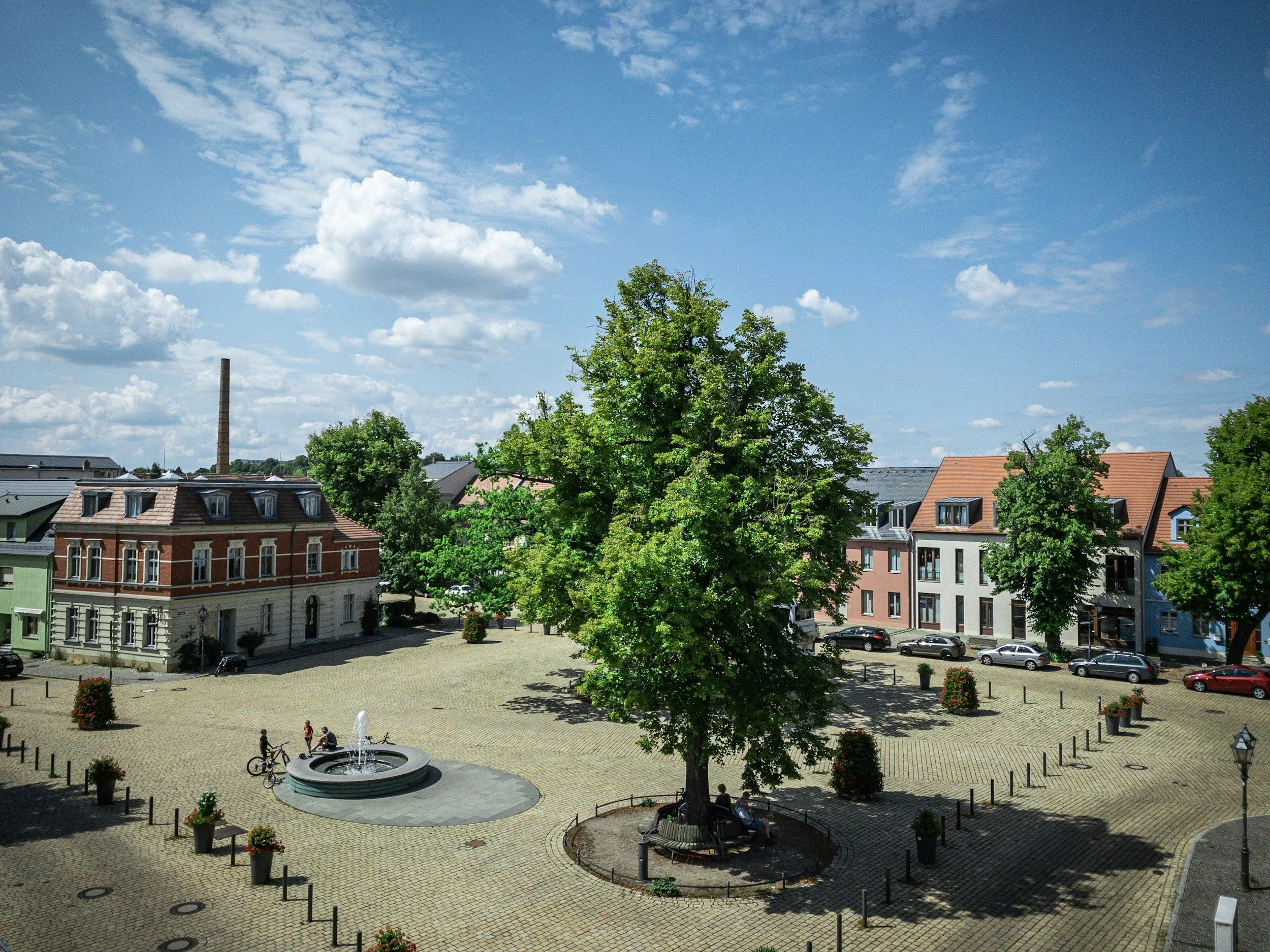 Stadtplatz mit Bäumen, Parkbank, Brunnen, bunt bemalten Häuser und einem blauen Himmel mit Wolken.