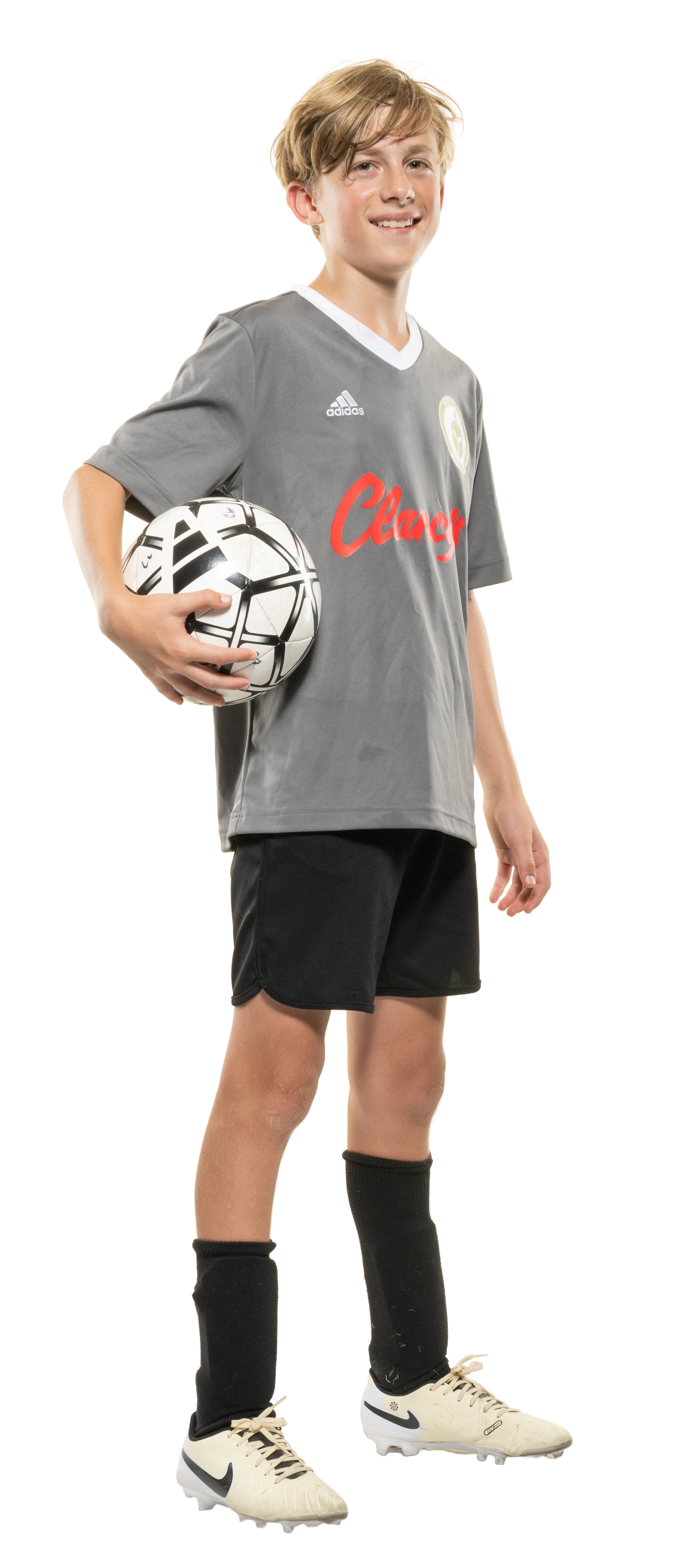 Young male soccer player holding a soccer ball, wearing a gray Adidas jersey with red text, black shorts, black knee-high socks, and white cleats.
