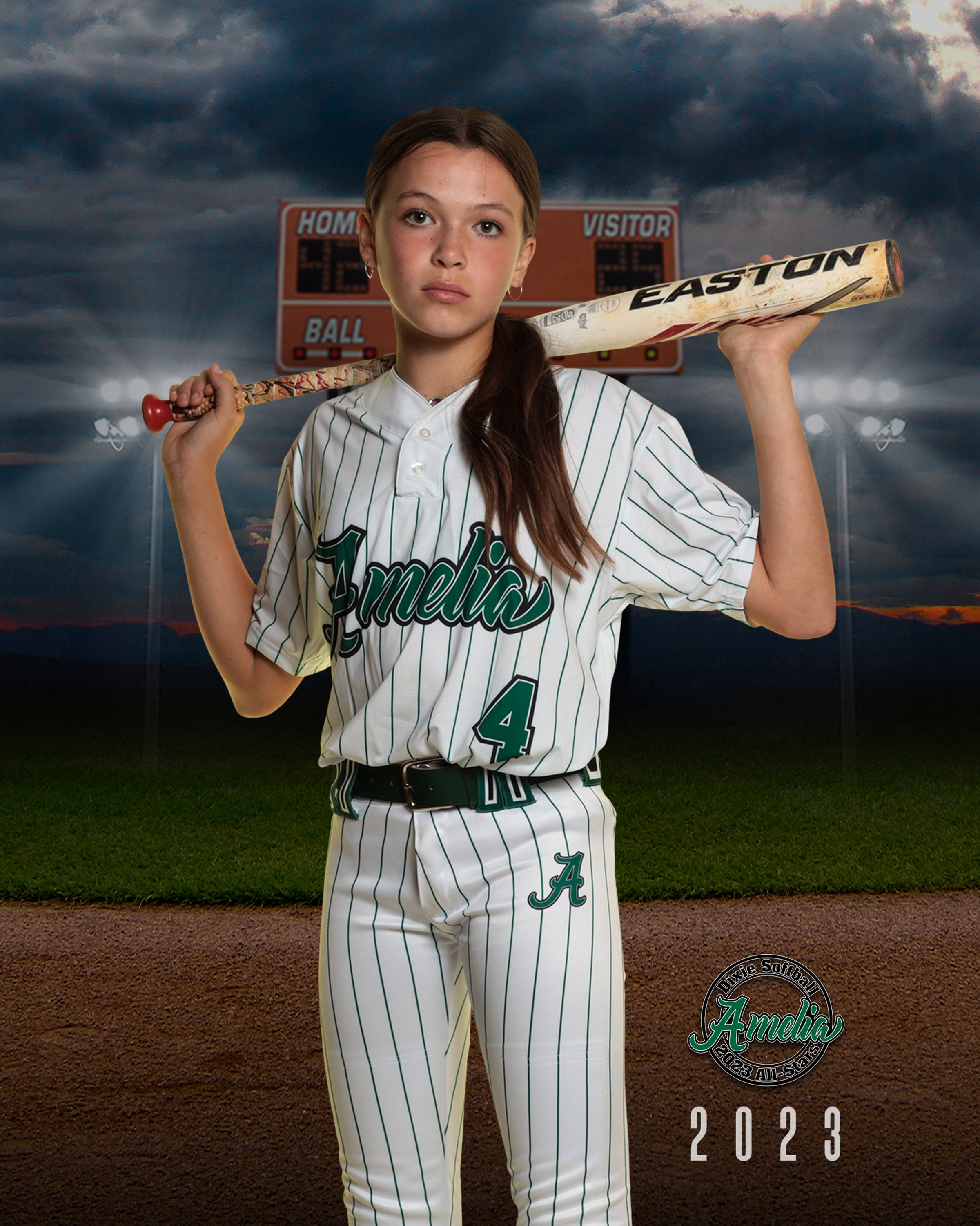 A young girl in a white baseball uniform with green pinstripes and the name 'Amelia' on the front, holding a baseball bat over her shoulder, standing on a baseball field at dusk with a scoreboard in the background.
