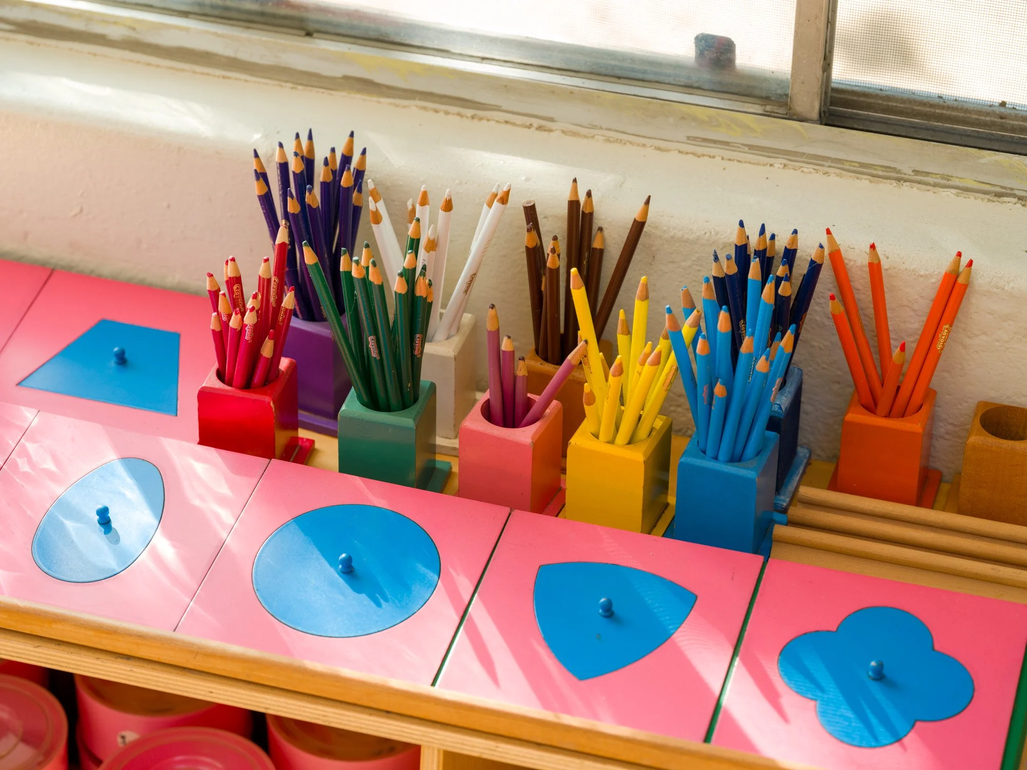 Colorful pencils organized in small square containers of various colors including red, purple, white, green, pink, yellow, blue, and orange, placed on a pink box with blue shapes on it, near a window.