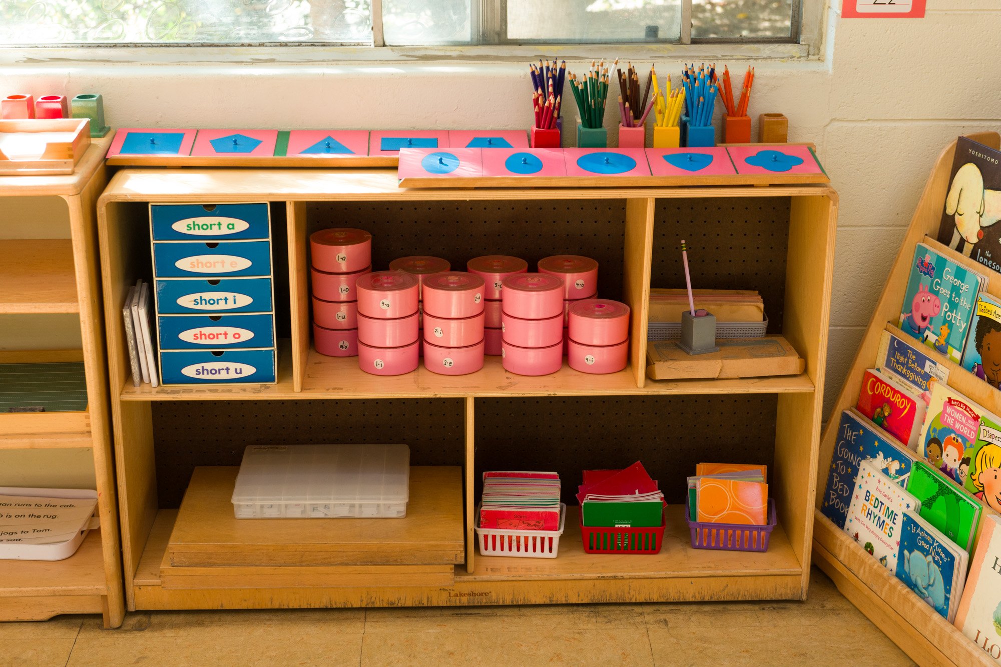 Wooden bookshelf with educational materials and books for children, including colored pencils, flashcards, and small storage containers.
