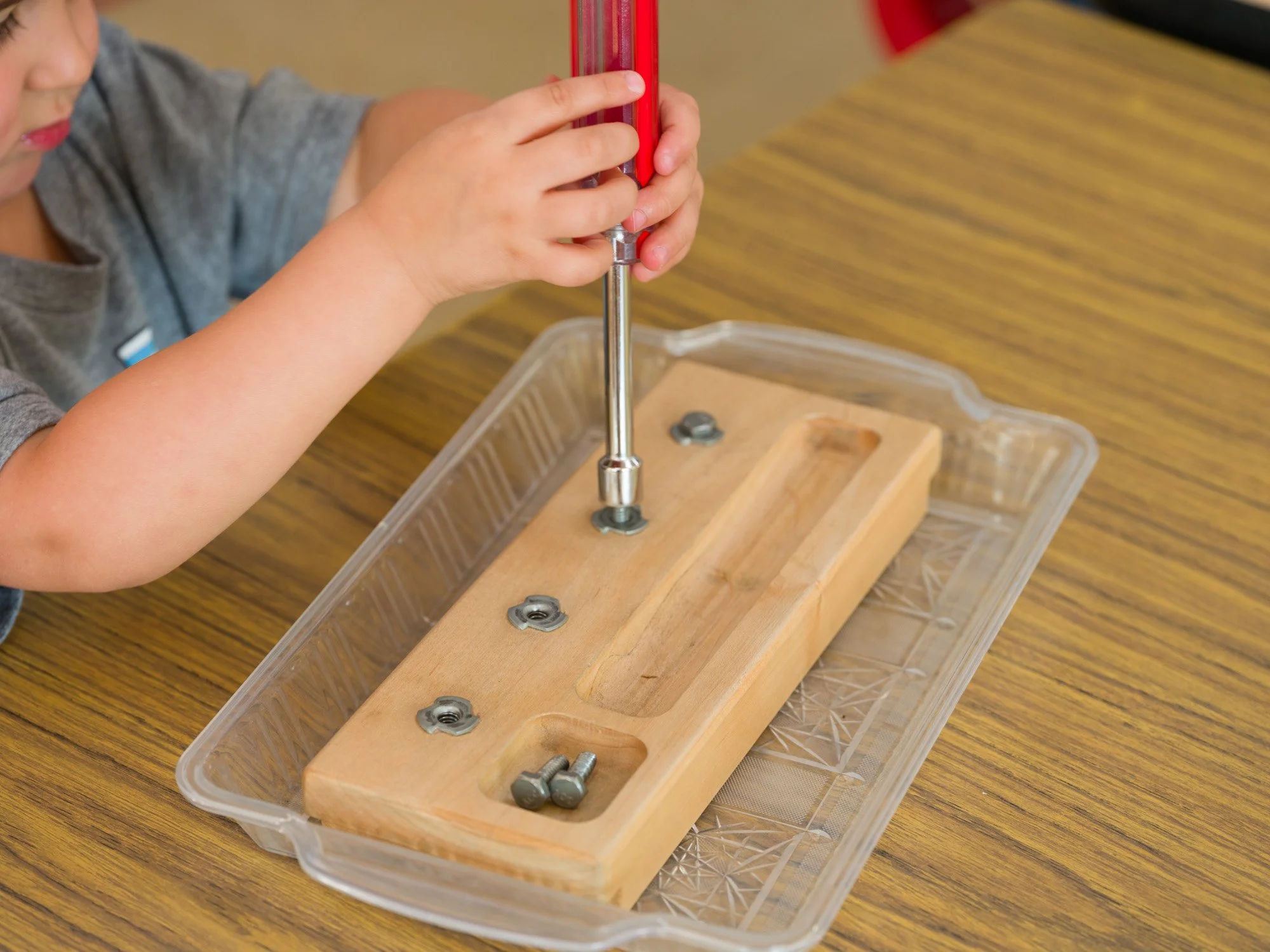Child using a red screwdriver to assemble a wooden board with metal screws and washers on a clear plastic tray.