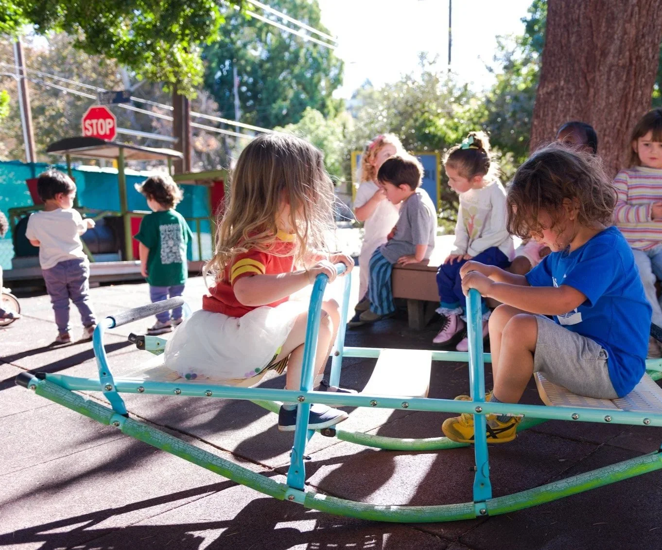 Children playing on a playground, some sitting on a seesaw and others standing or sitting nearby, with trees and a stop sign in the background.