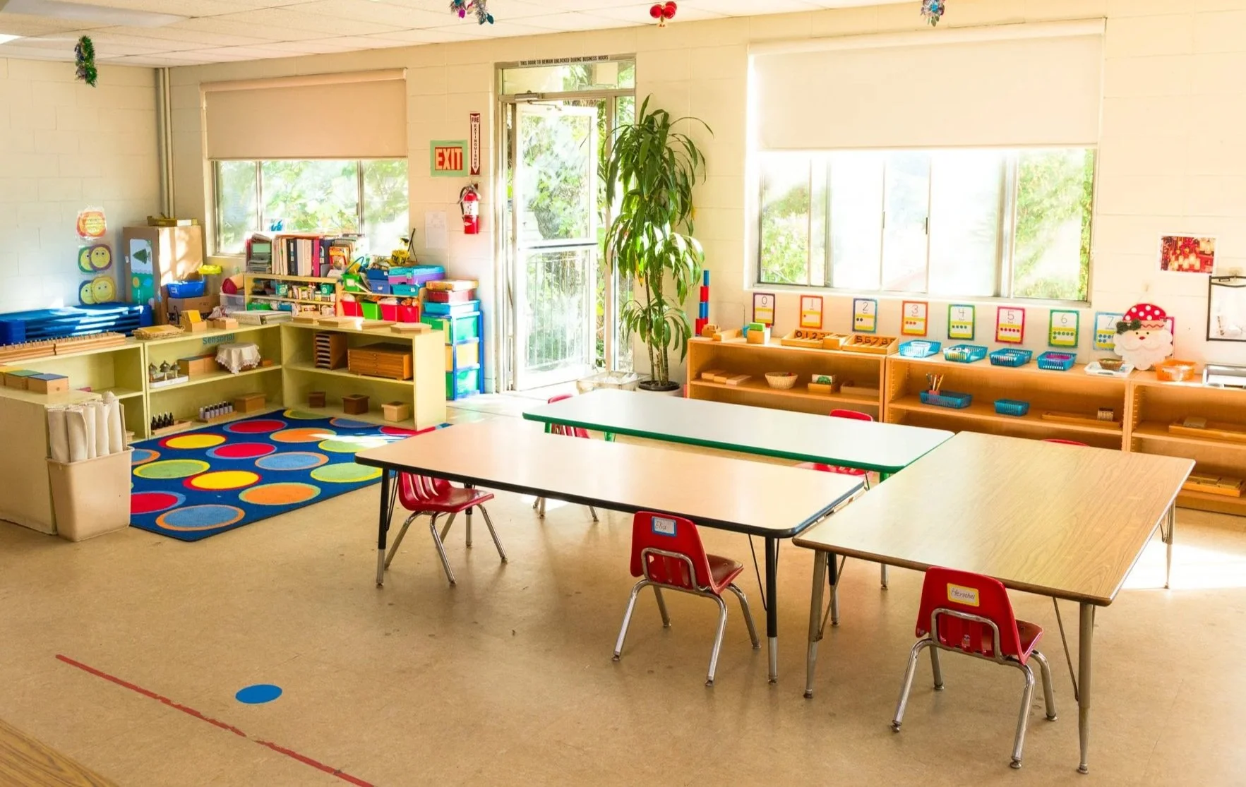 Bright and colorful preschool classroom with tables, chairs, educational supplies, window, and door.