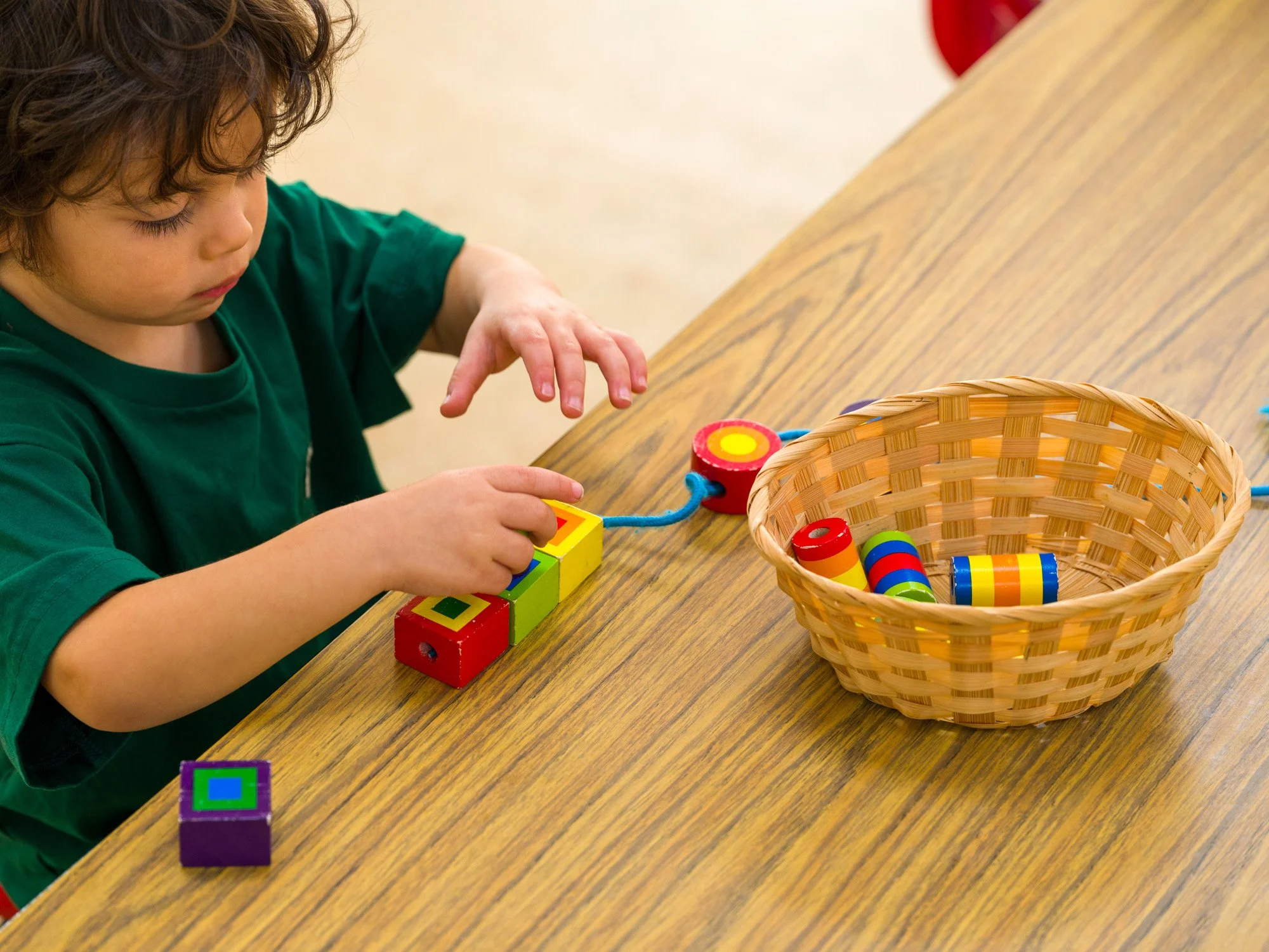 A young child with curly hair and a green shirt is playing with colorful stacking blocks on a wooden table. There is a small basket containing more stacking blocks nearby.