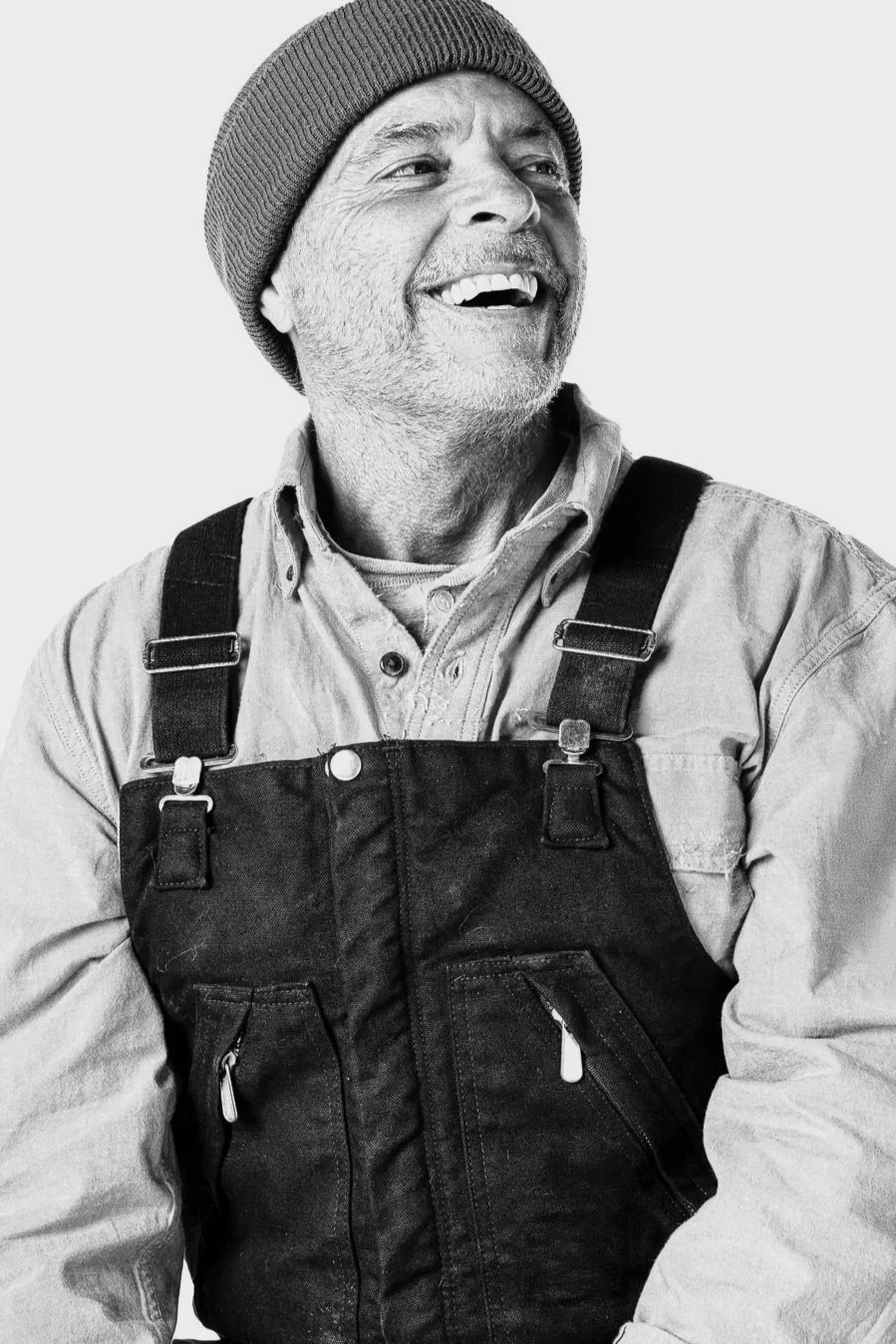 A smiling older man wearing a beanie, a button-up shirt, and black overalls, looking away from the camera.