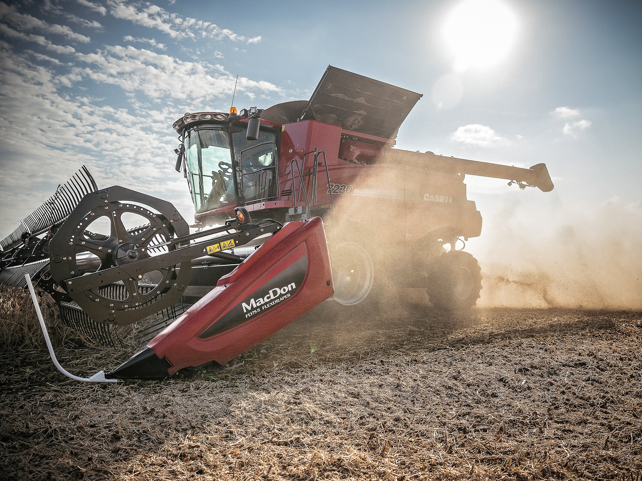 A large red and black combine harvester working in a dry field, kicking up dust under a partly cloudy sky.