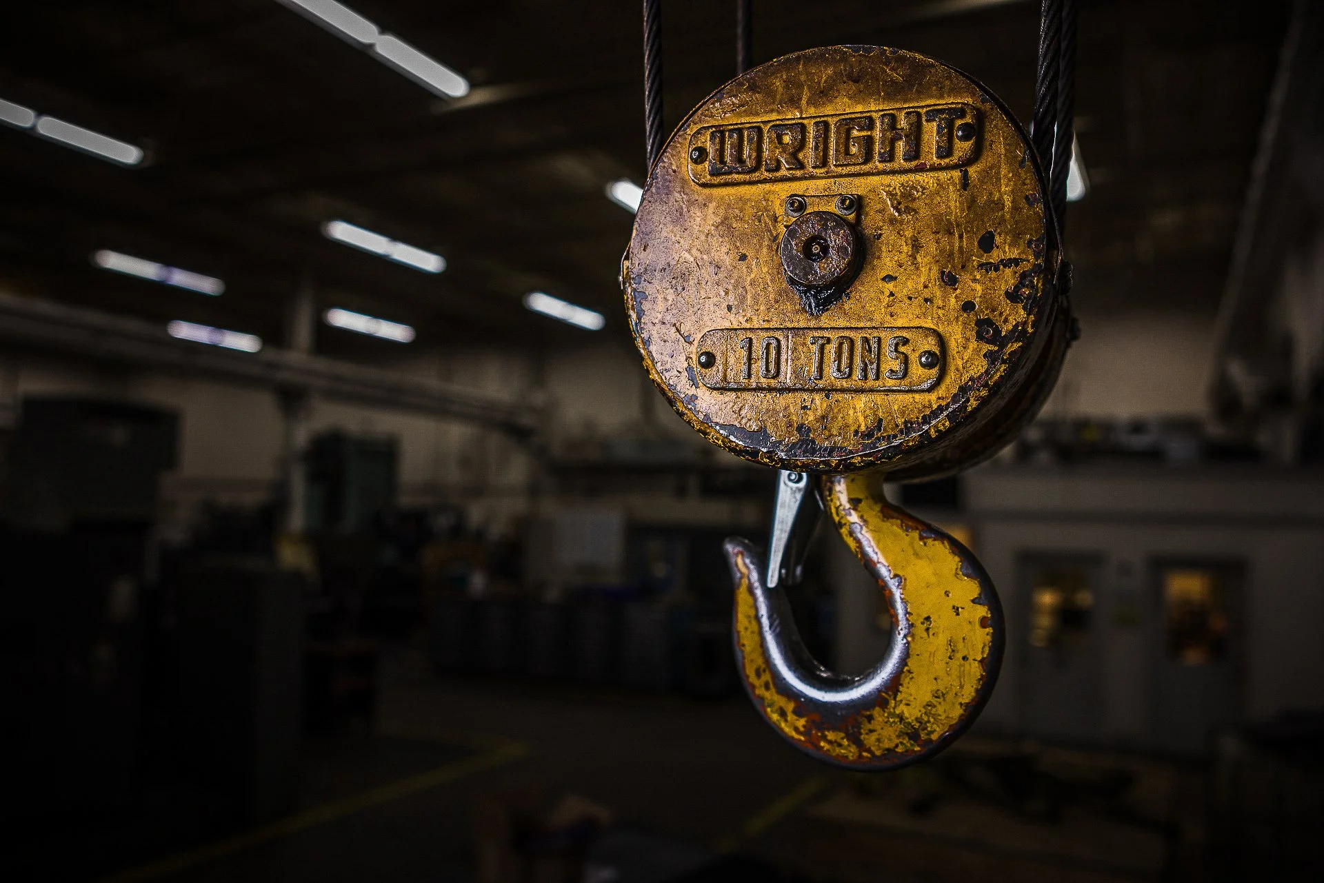 Close-up of a yellow overhead crane hook with the words 'WRIGHT' and '10 TONS' embossed on it, in an industrial warehouse.