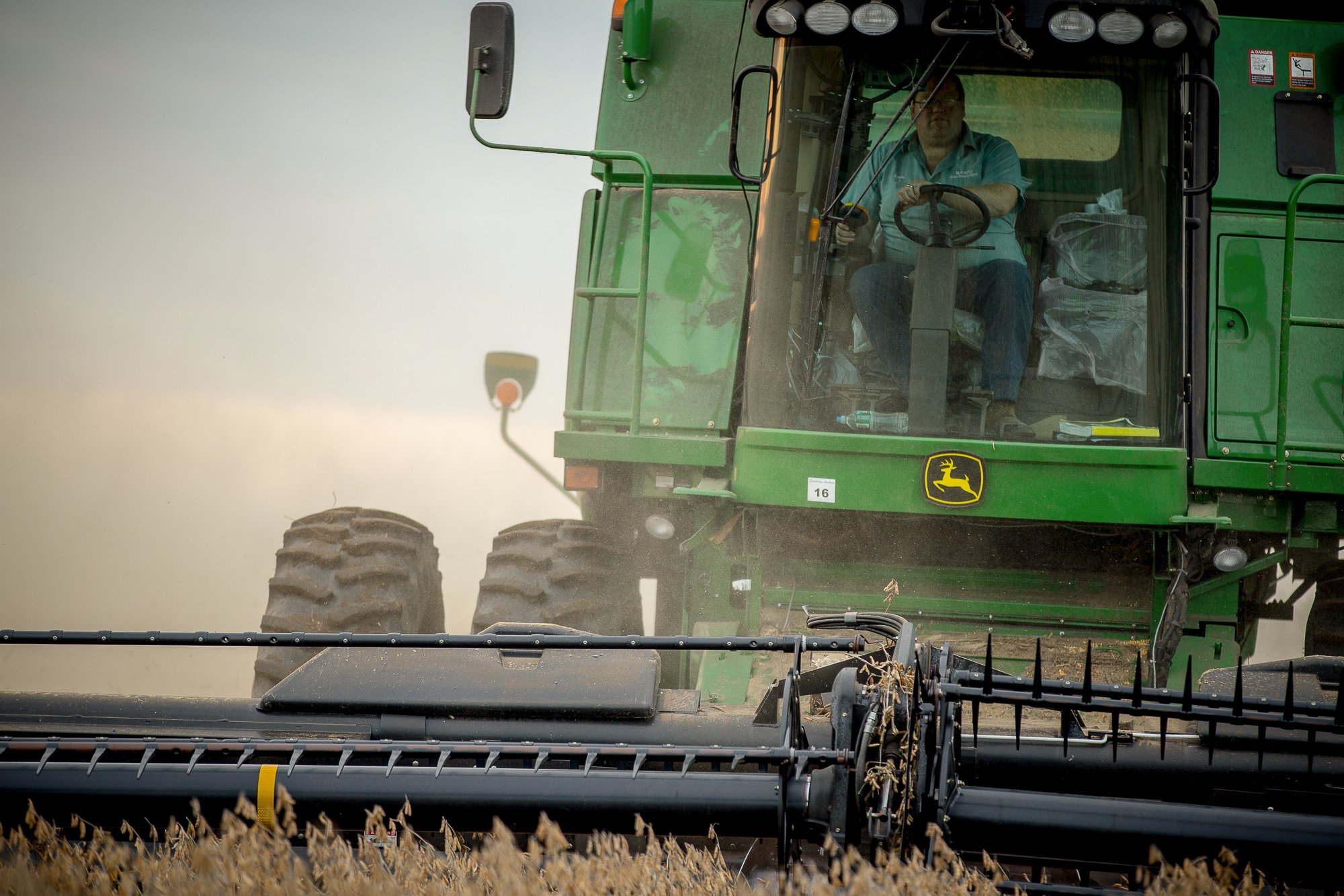 A man driving a green John Deere combine harvester in a field, harvesting crops on a dusty day.