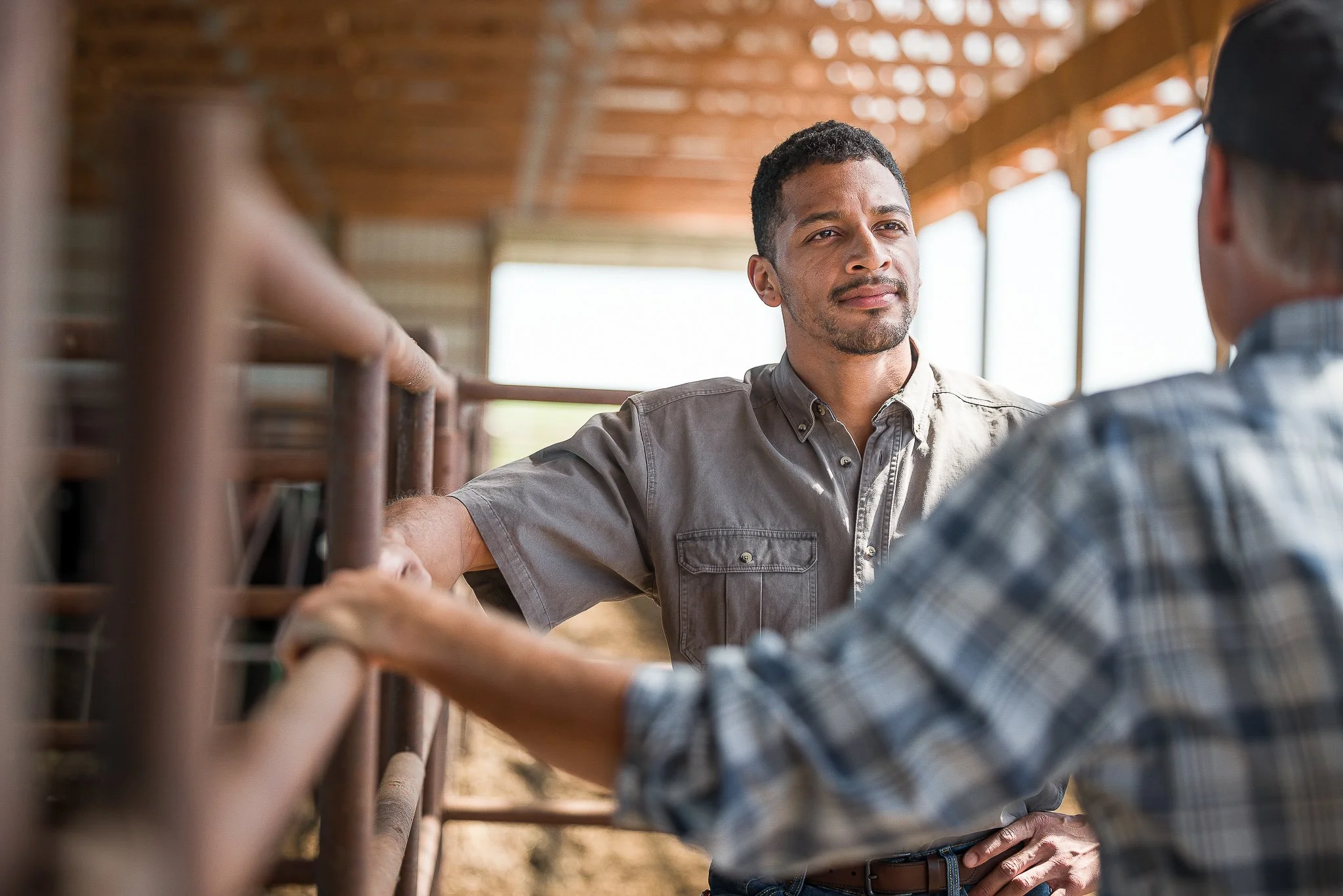 Two men having a conversation inside a barn or stable, one man with a beard and wearing a gray shirt, the other man with glasses and wearing a plaid shirt.
