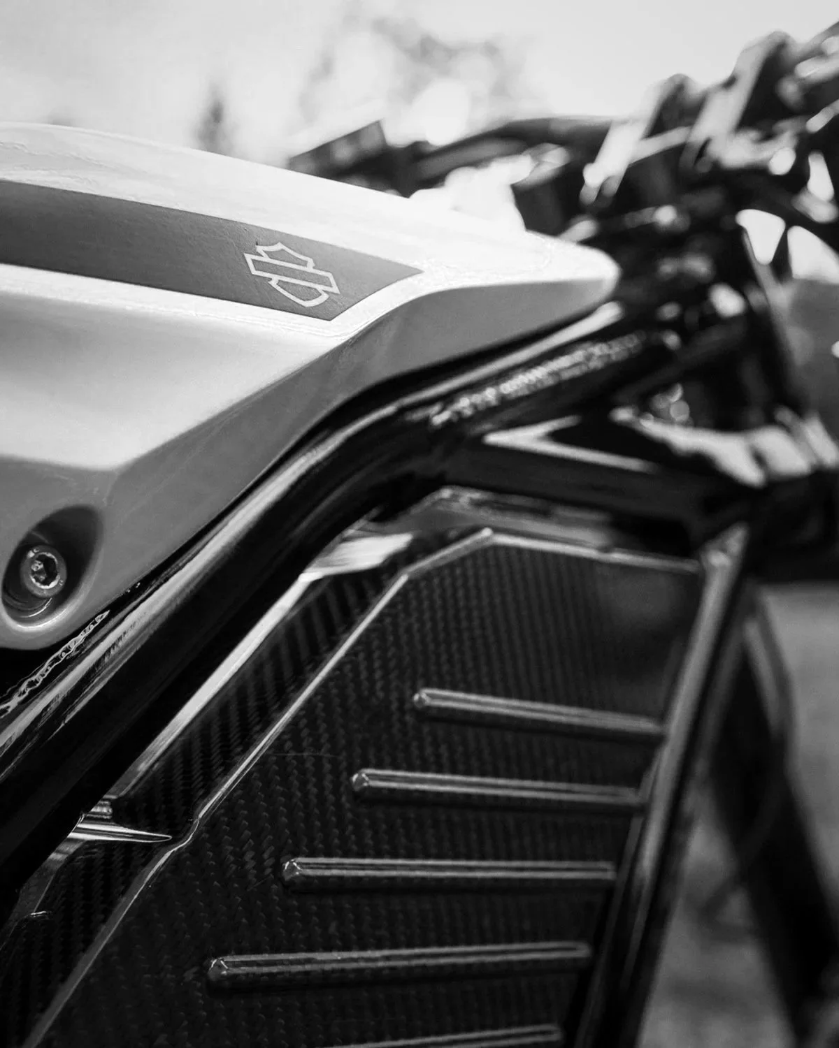 Close-up black and white photo of a motorcycle's front section, showing part of the fuel tank with a Harley-Davidson logo and a carbon fiber grille.