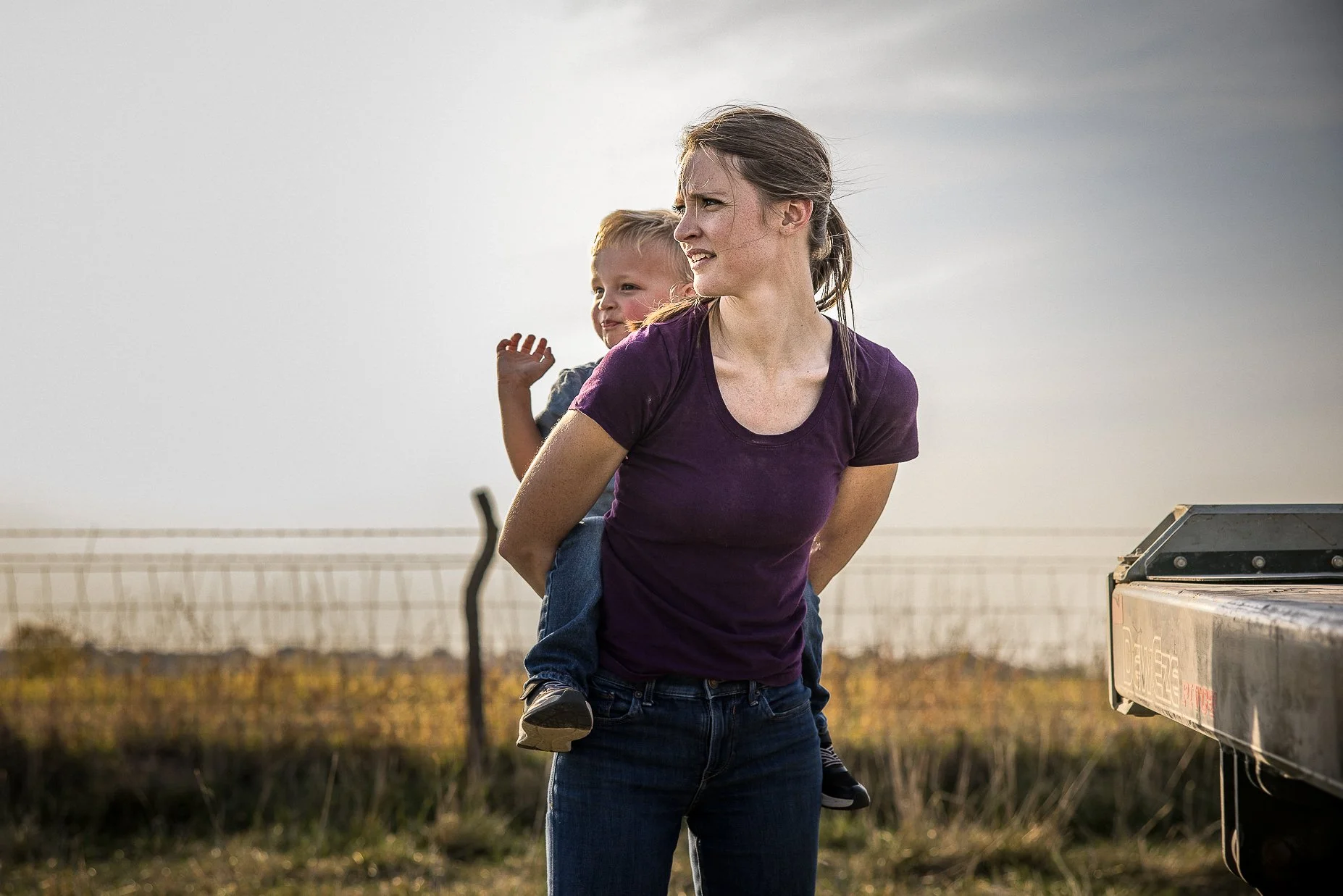 A woman carrying a young child on her back outdoors during late afternoon or early evening, with an open field and fence in the background.