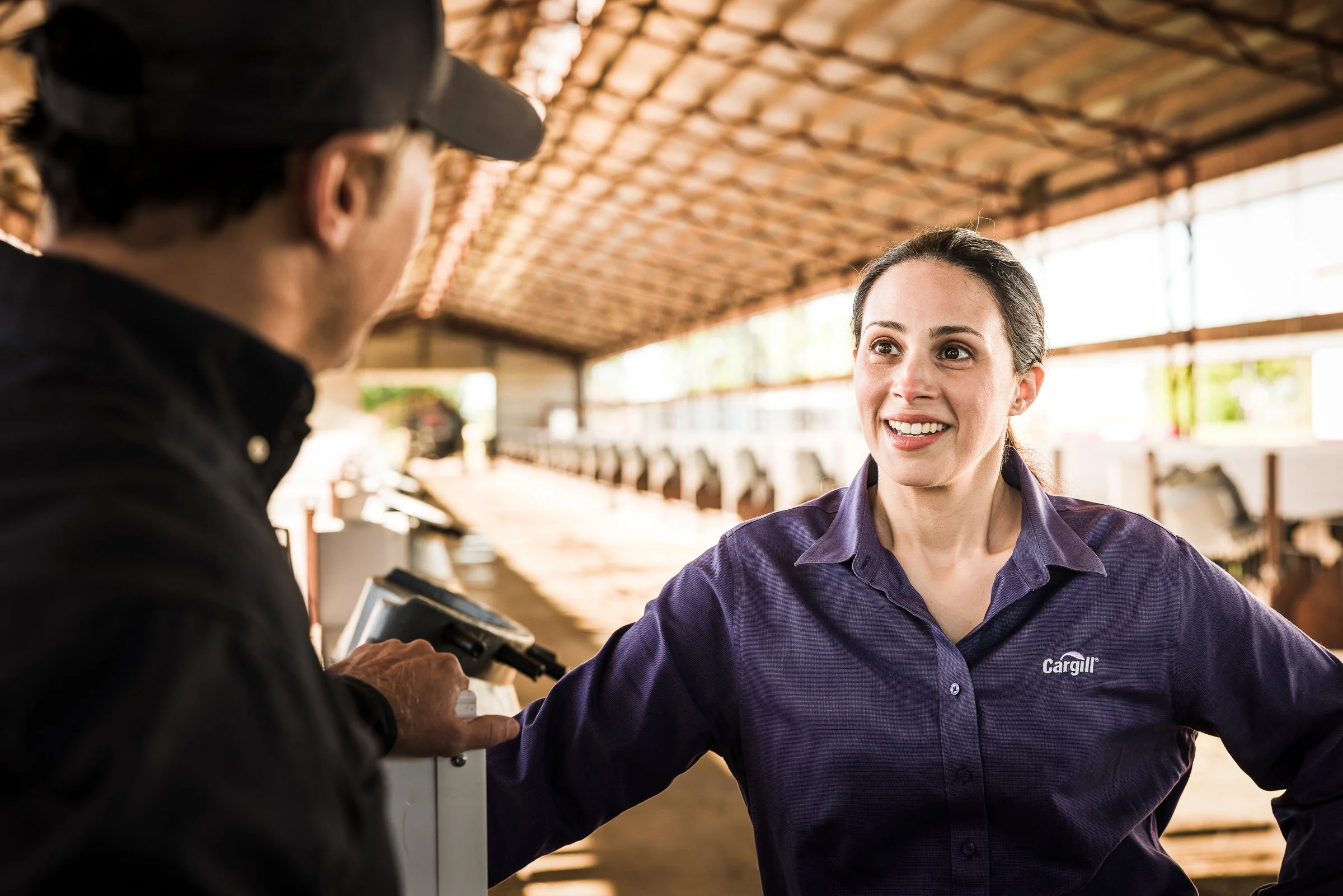 A woman in a purple shirt labeled 'Cargill' smiling and talking to a man in a black shirt in a barn or livestock facility.