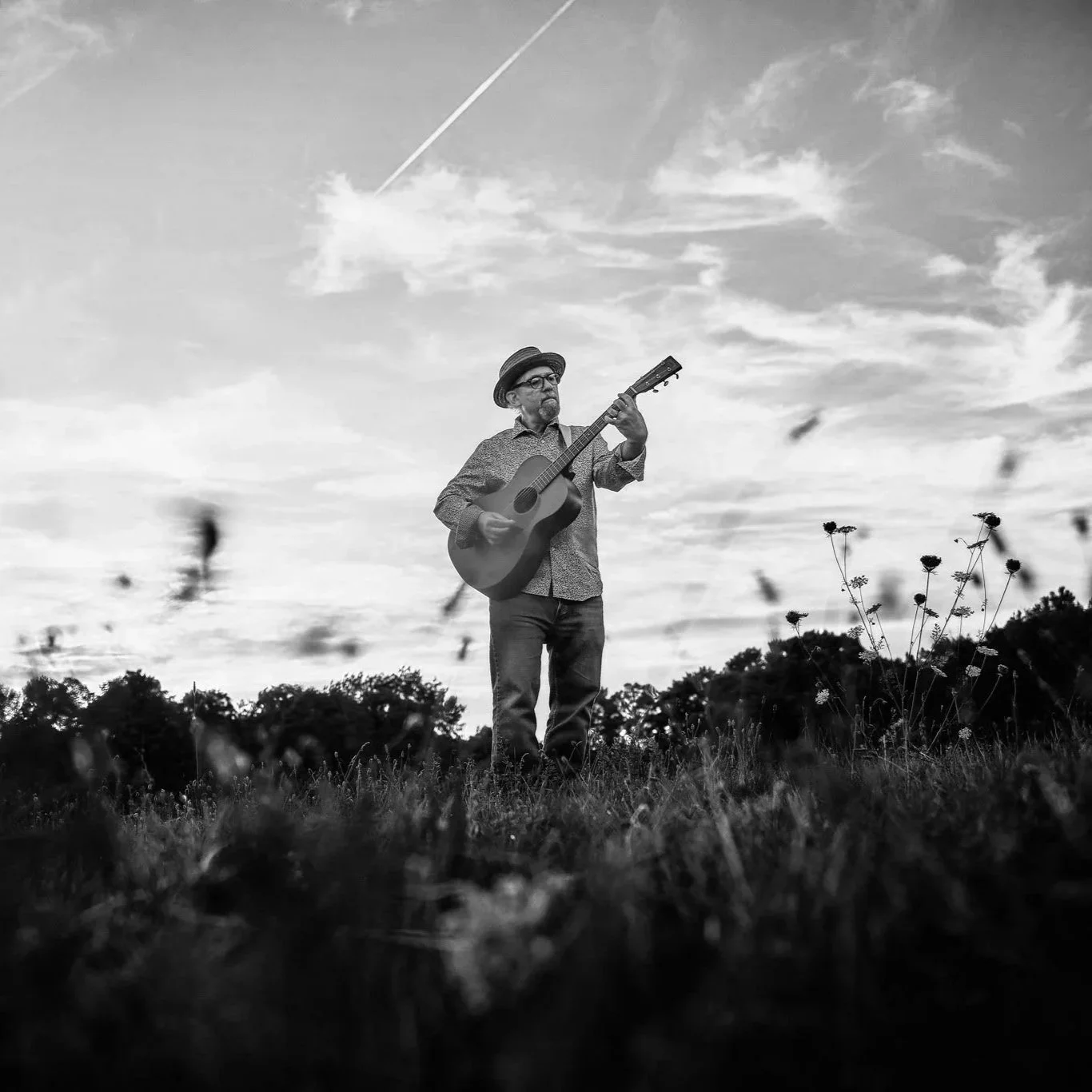A man wearing a hat and glasses playing an acoustic guitar outdoors in a field during the daytime, black and white photo, with clouds and contrail in the sky.