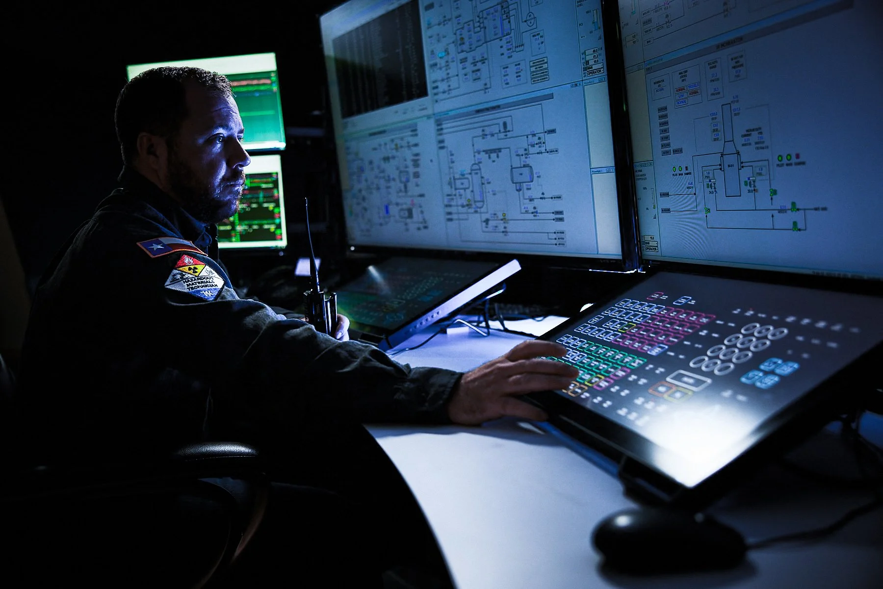A person in military uniform operating multiple large monitors displaying technical diagrams and data in a dark room.