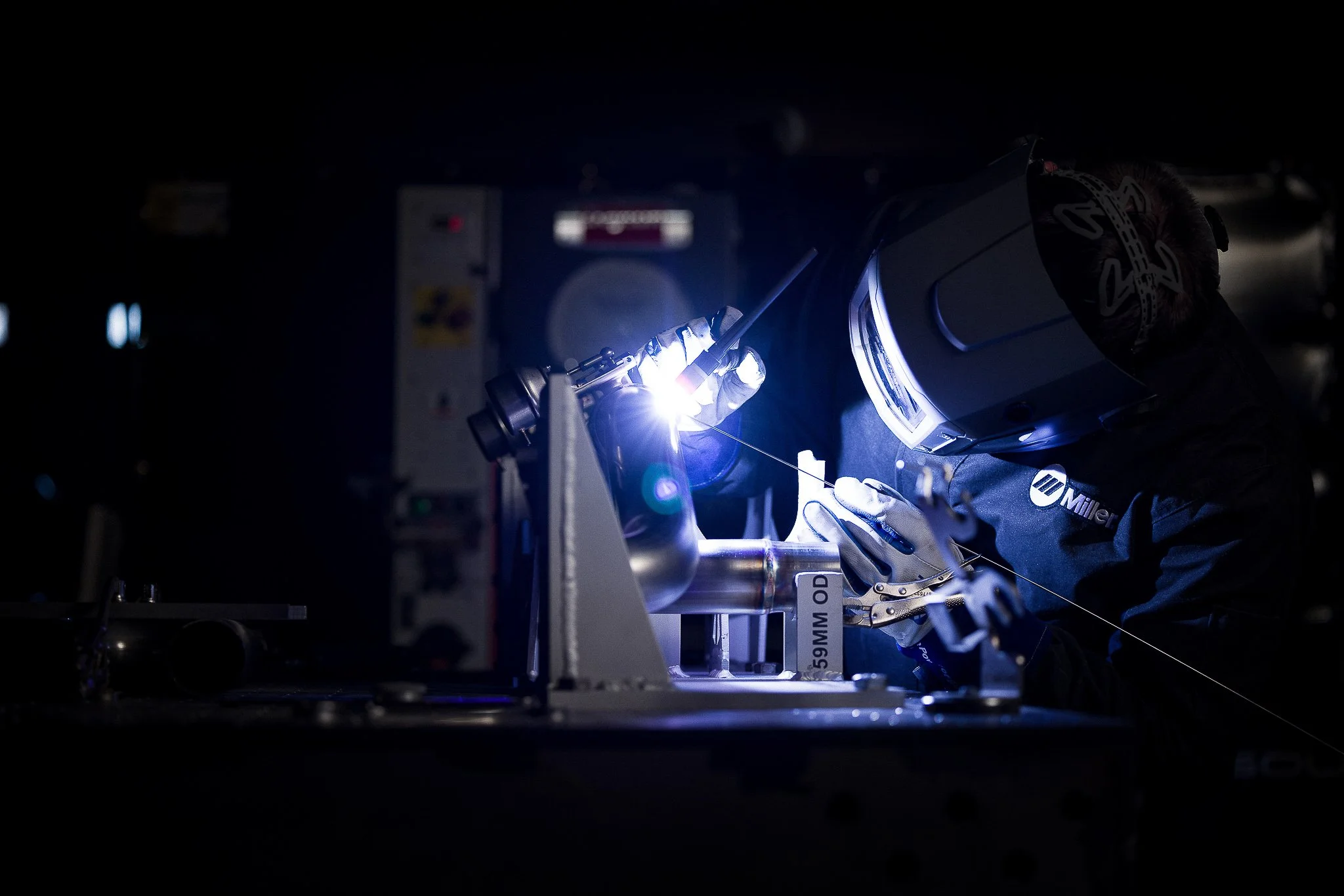 A person in protective gear, including a welding helmet, working on a metal component with a torch in a dark environment.