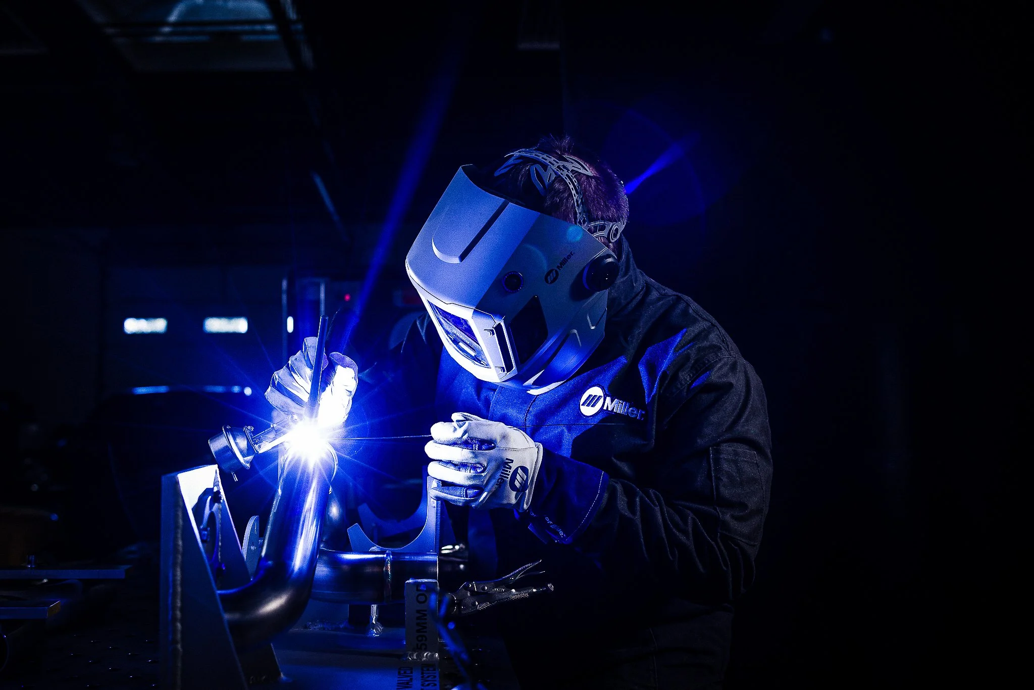 A welder wearing a helmet and gloves welding metal with bright sparks and blue light in a dark industrial setting.
