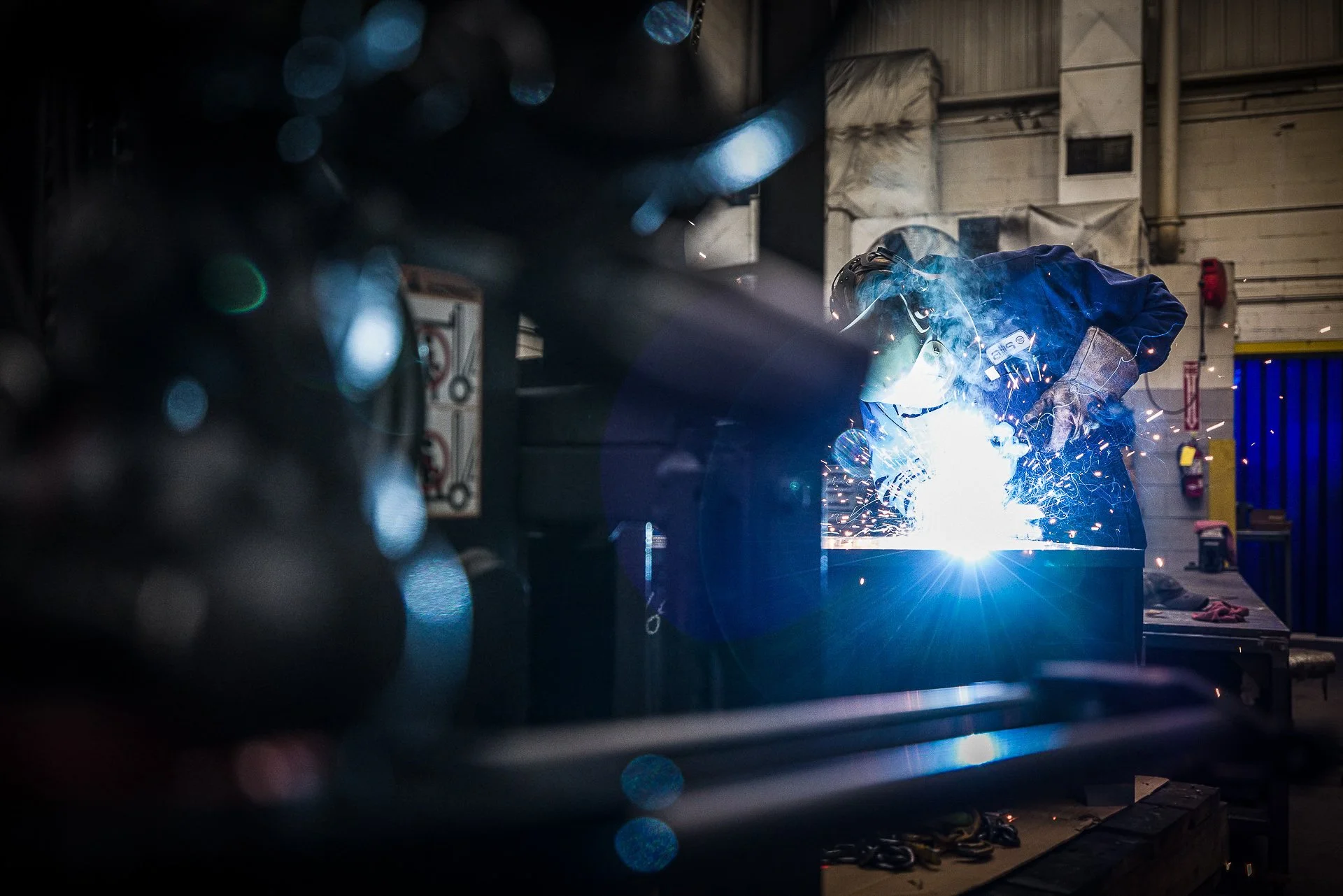 Welder wearing protective gear welding metal in an industrial workshop with sparks flying.