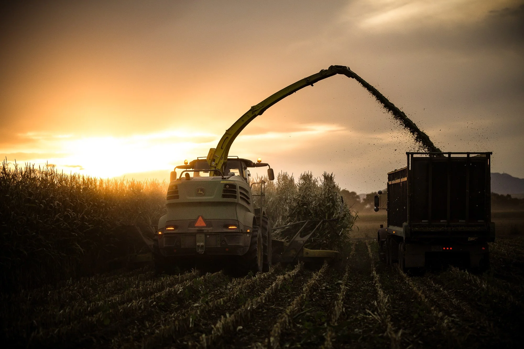 A tractor with a large arm unloading harvested crops into a truck during sunset over farmland.