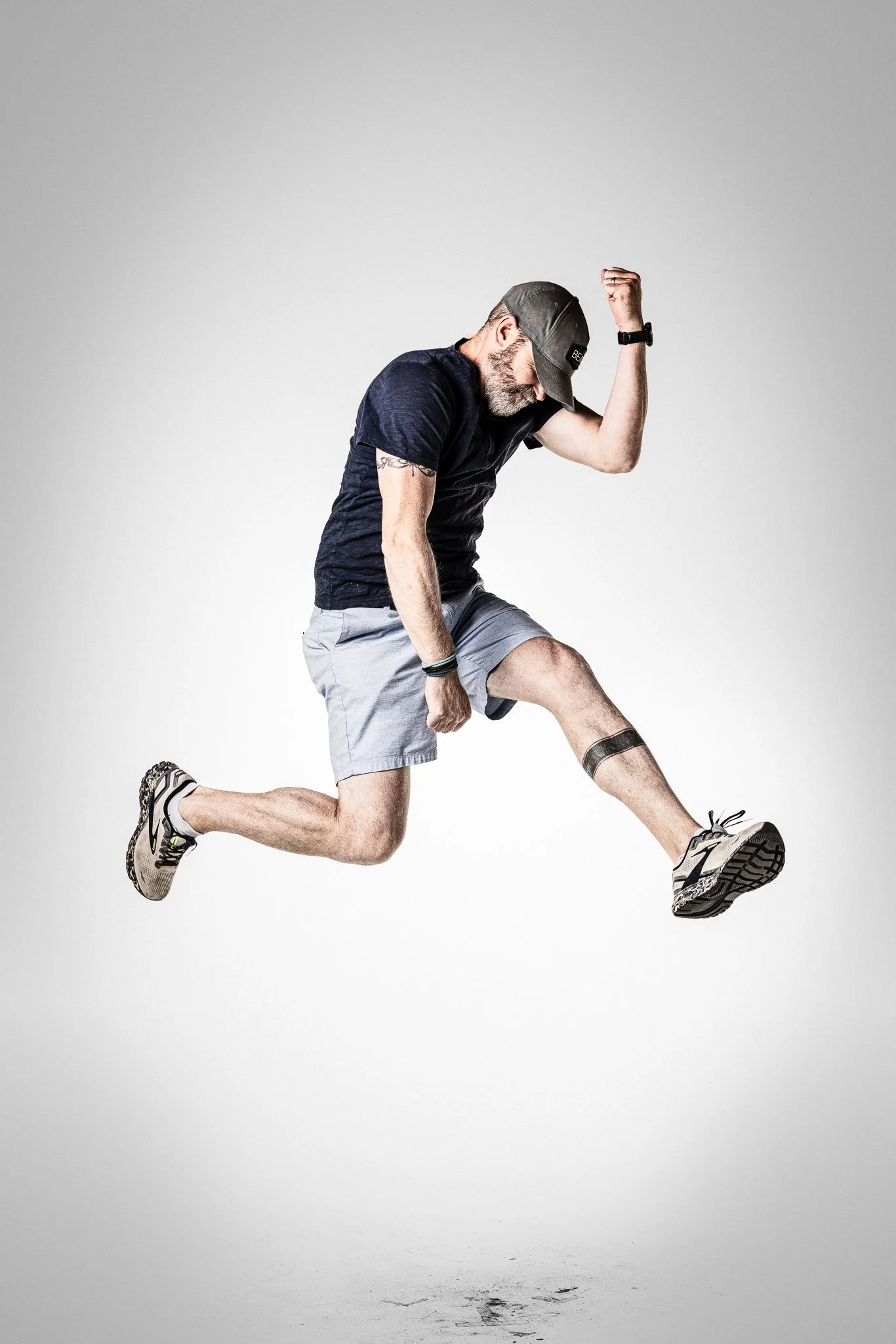 A man wearing a cap, black t-shirt, and shorts jumping in the air with a joyful expression, against a plain gray background.