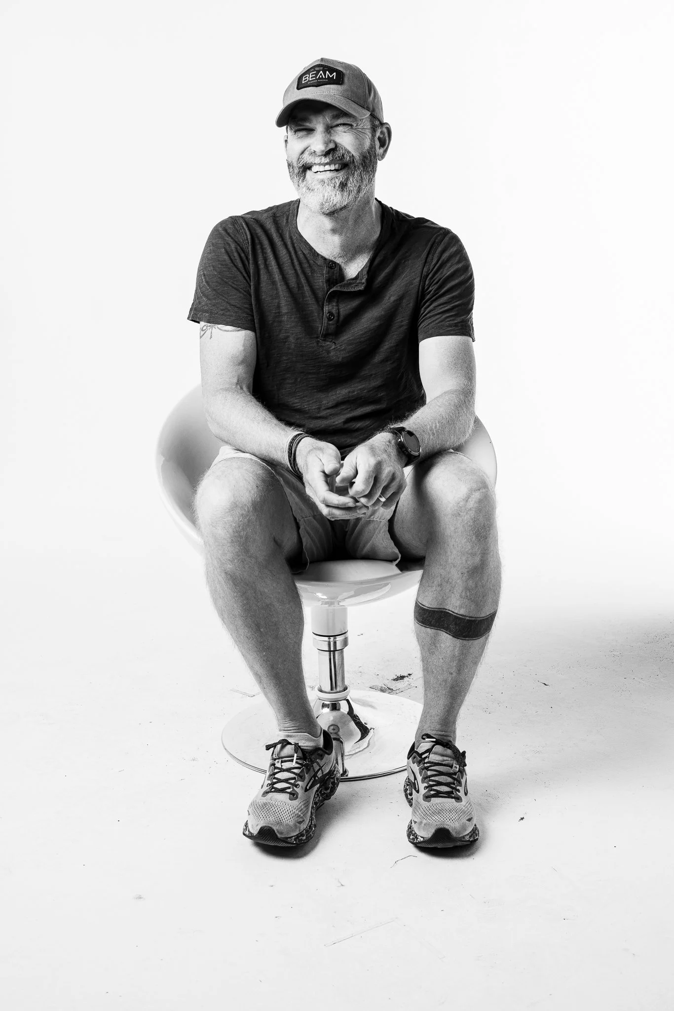 A man wearing a baseball cap, a black T-shirt, shorts, and sneakers is sitting on a modern chair against a white background, smiling with a beard and a tattoo on his right arm.