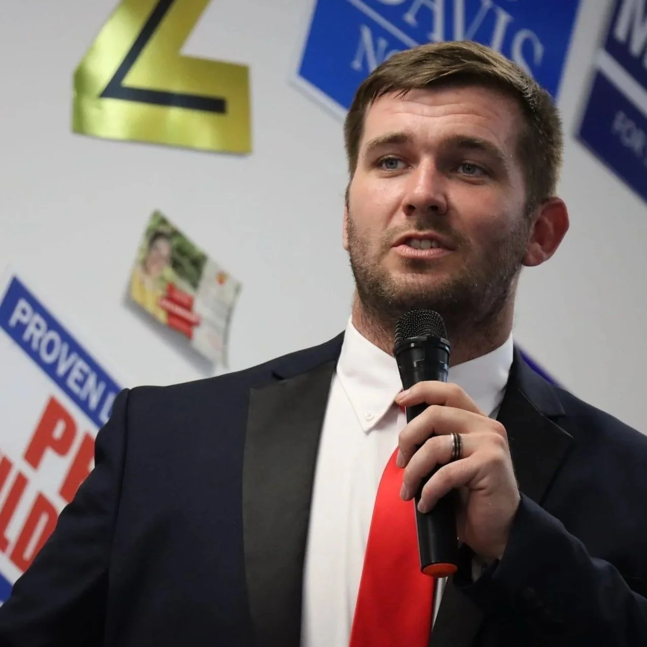 A young man in a suit and red tie holding a microphone while speaking at an event with political signs in the background.