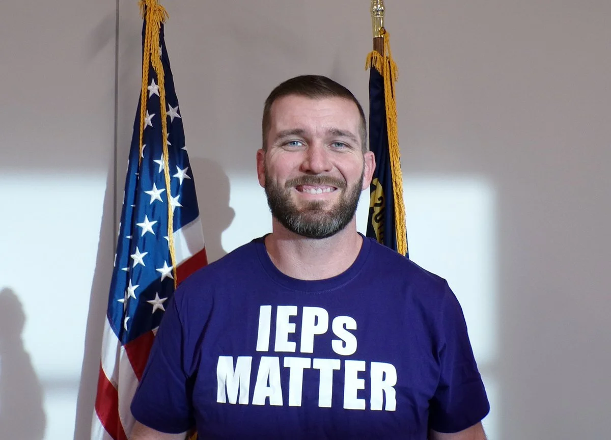 A smiling man with a beard stands in front of American flags, wearing a blue T-shirt that says 'IEPs MATTER'.