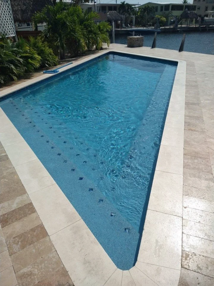 A rectangular swimming pool with clear blue water, surrounded by a tiled deck and lush greenery near a waterfront.