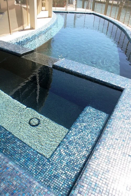 Indoor swimming pool with tiled edges and a hot tub, located in a sunlit room with large windows.