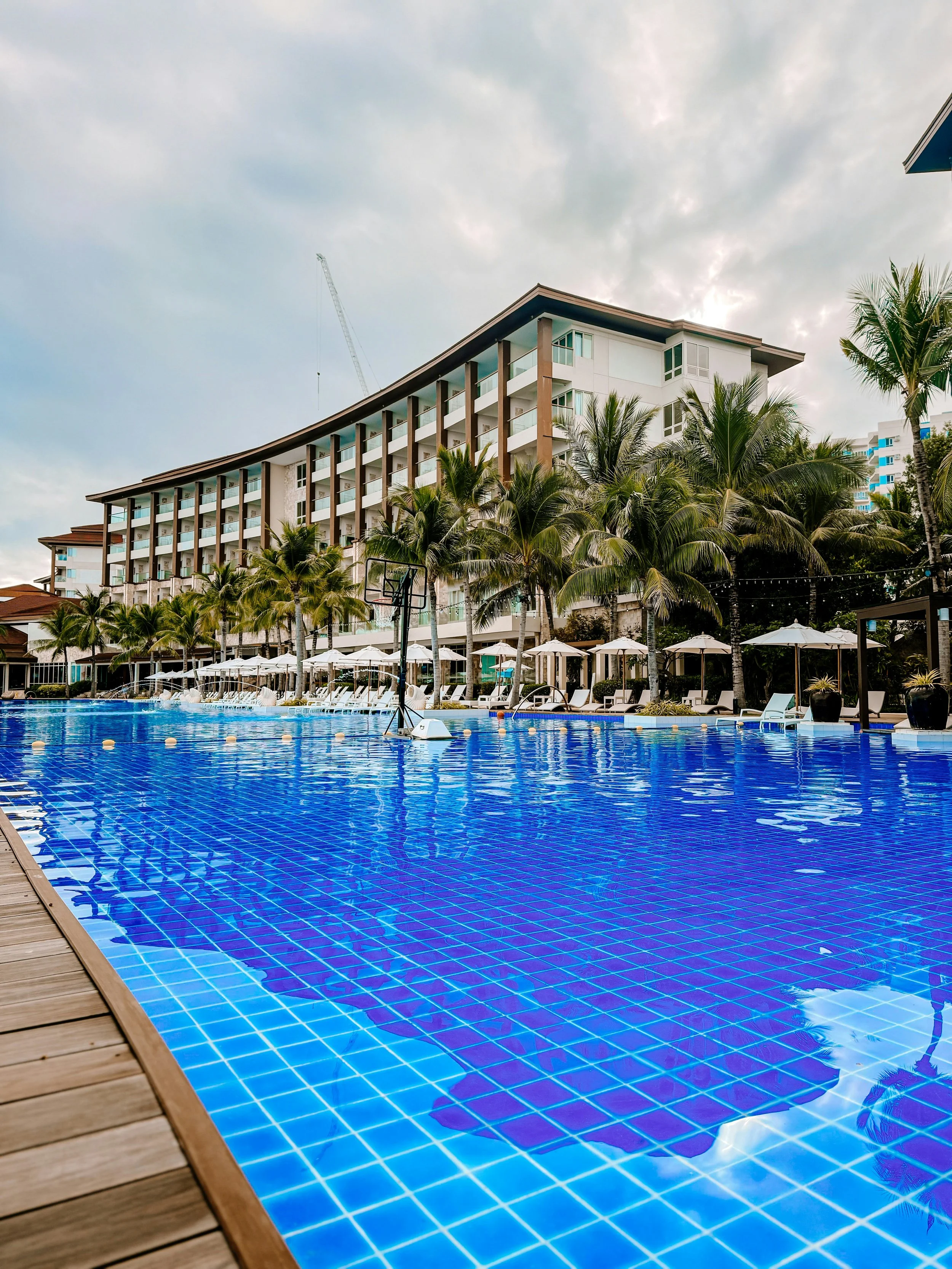 A swimming pool with blue tiles at a resort, surrounded by lounge chairs, umbrellas, and palm trees, with a large hotel building in the background.