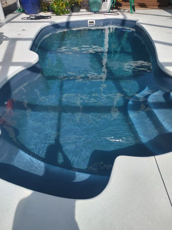 An empty outdoor hot tub with clear water, surrounded by a white concrete deck, with potted plants and gardening tools in the background.