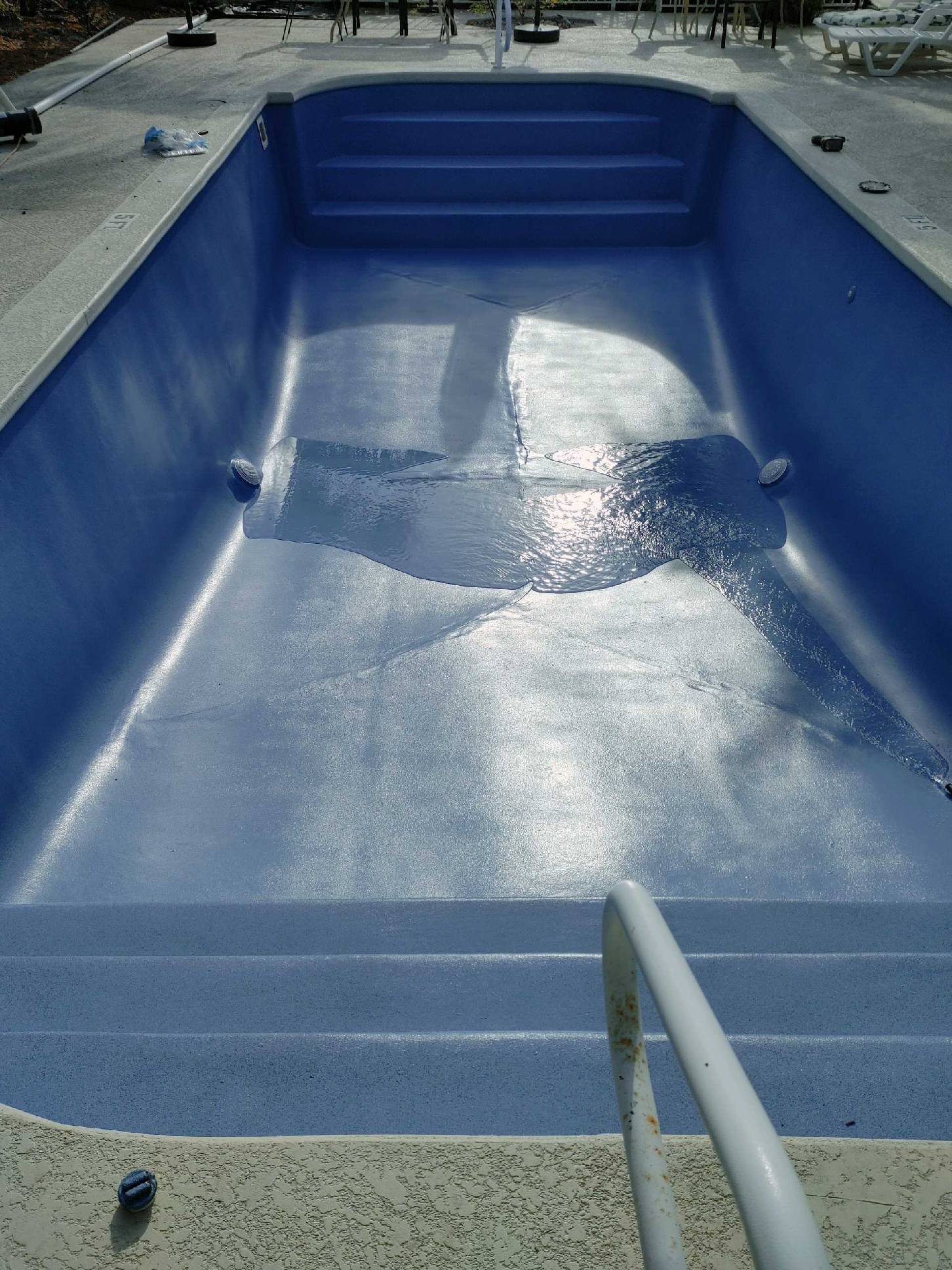 Empty swimming pool with a dolphin-shaped repair patch on the floor, surrounded by poolside lounge chairs and umbrellas.