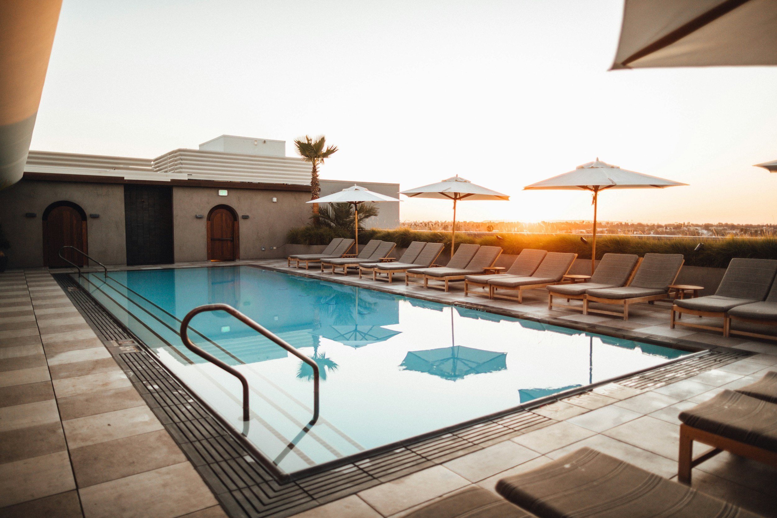 An outdoor swimming pool area at sunset with lounge chairs, white umbrellas, and a city skyline in the distance.