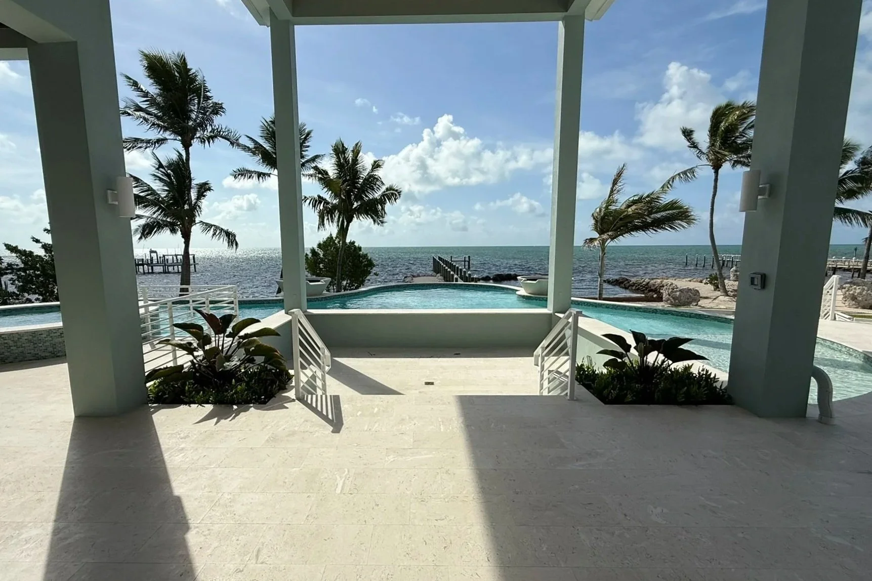 View of a luxurious beachfront patio with a pool, overlooking the ocean, with palm trees and a pier in the background.