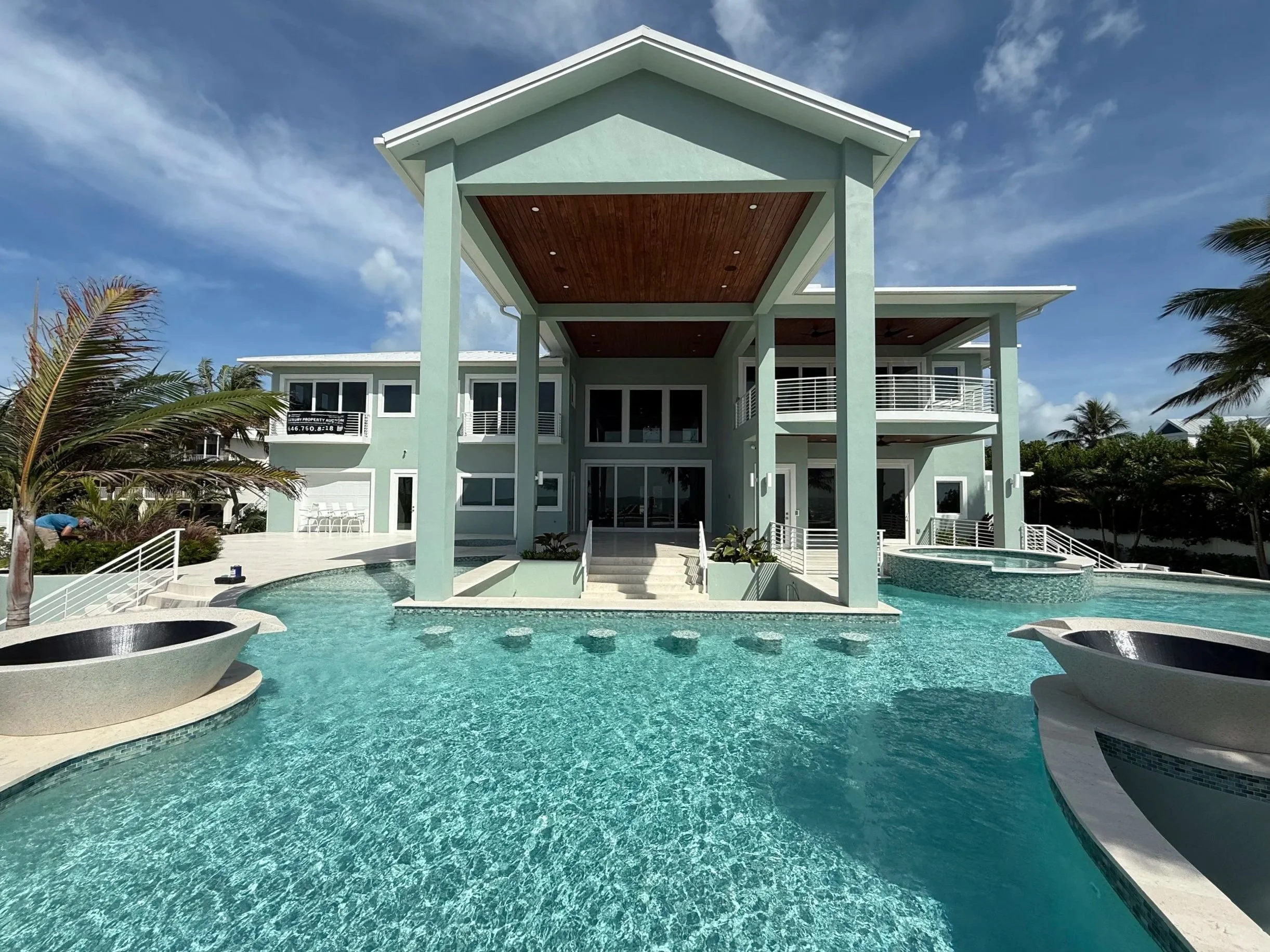 A luxurious modern house with a large swimming pool in the front, surrounded by tropical trees. The house is light green with multiple balconies and large glass windows, under a blue sky with few clouds.