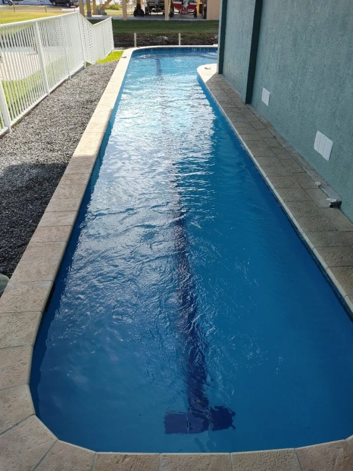 A narrow backyard pool with blue water, surrounded by beige tile and white fence on one side, next to a light blue house wall.