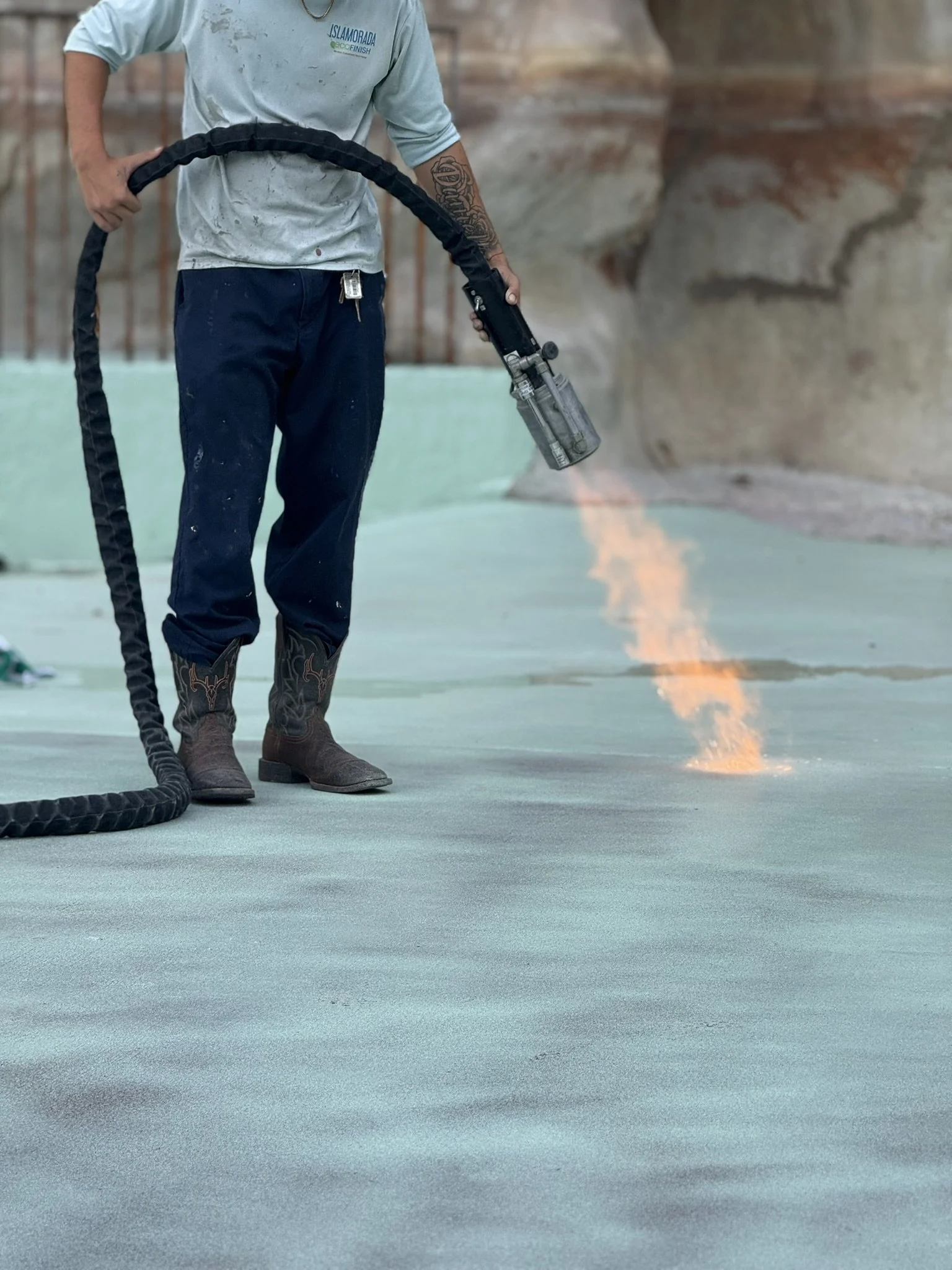 Person wearing jeans, boots, and a T-shirt using a blowtorch to melt asphalt on a roof surface.
